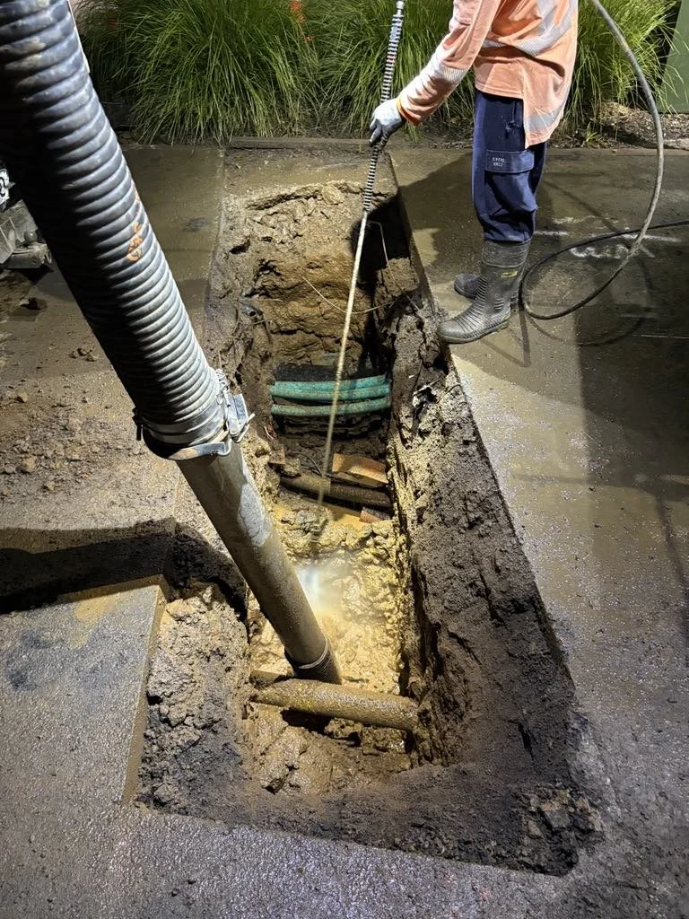 A person standing near a trench performing maintenance work. The trench contains multiple pipes and is being vacuumed or cleaned with a large hose. The person is wearing waterproof boots and a safety vest. The area around the trench is wet and muddy.