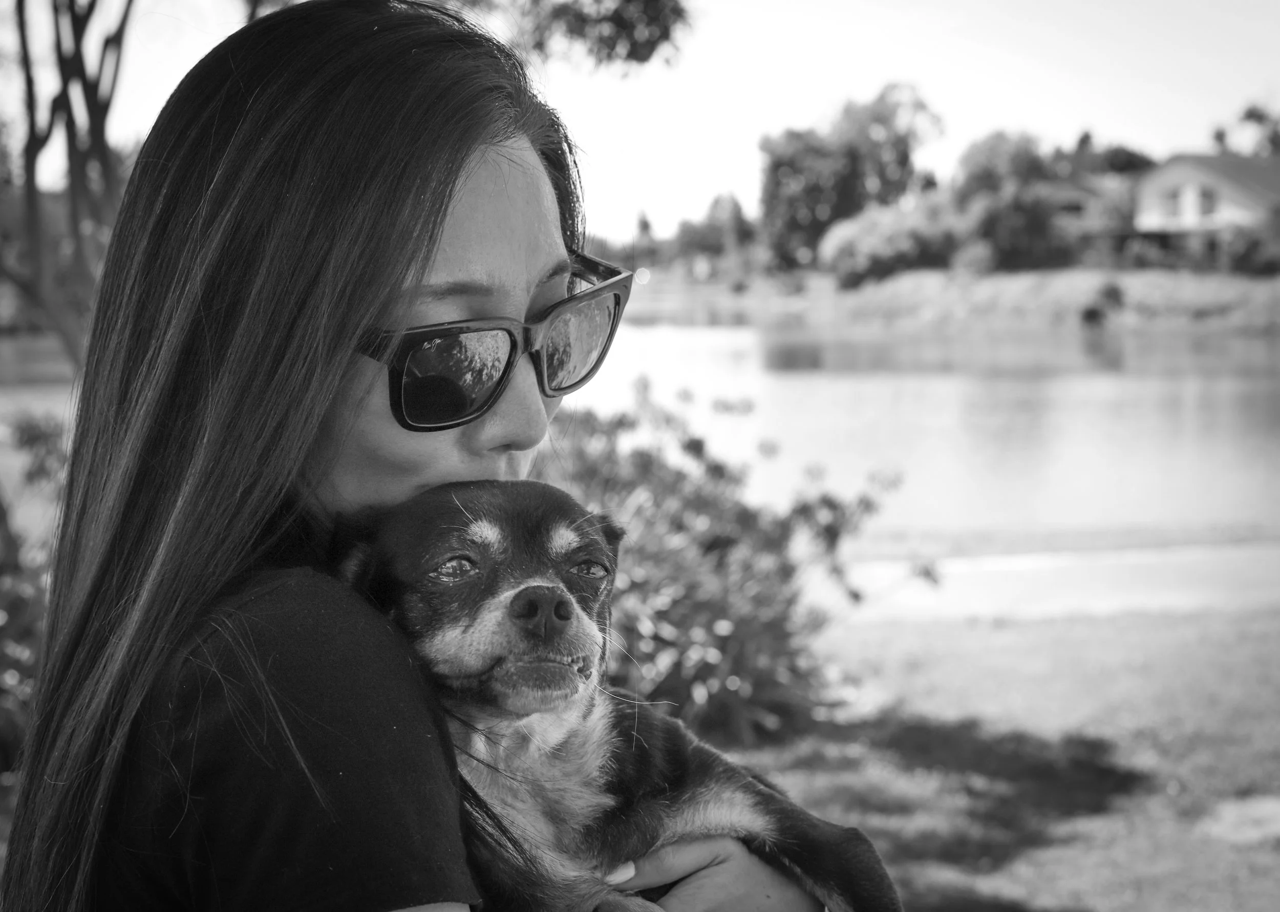 A woman wearing glasses is holding a small dog close to her chest near a river, with trees and houses in the background.