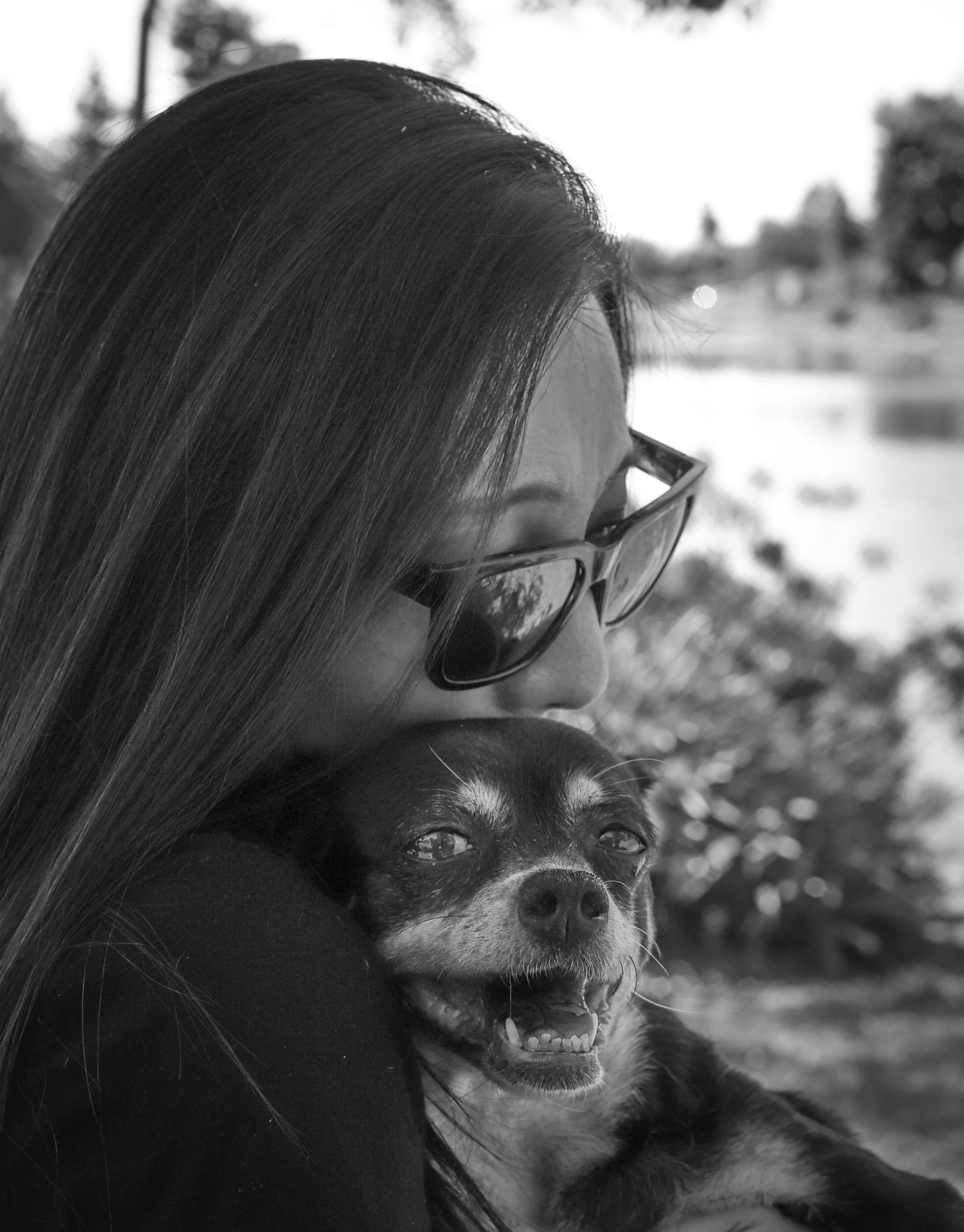 A woman with long hair and sunglasses hugging a small, smiling dog outdoors.