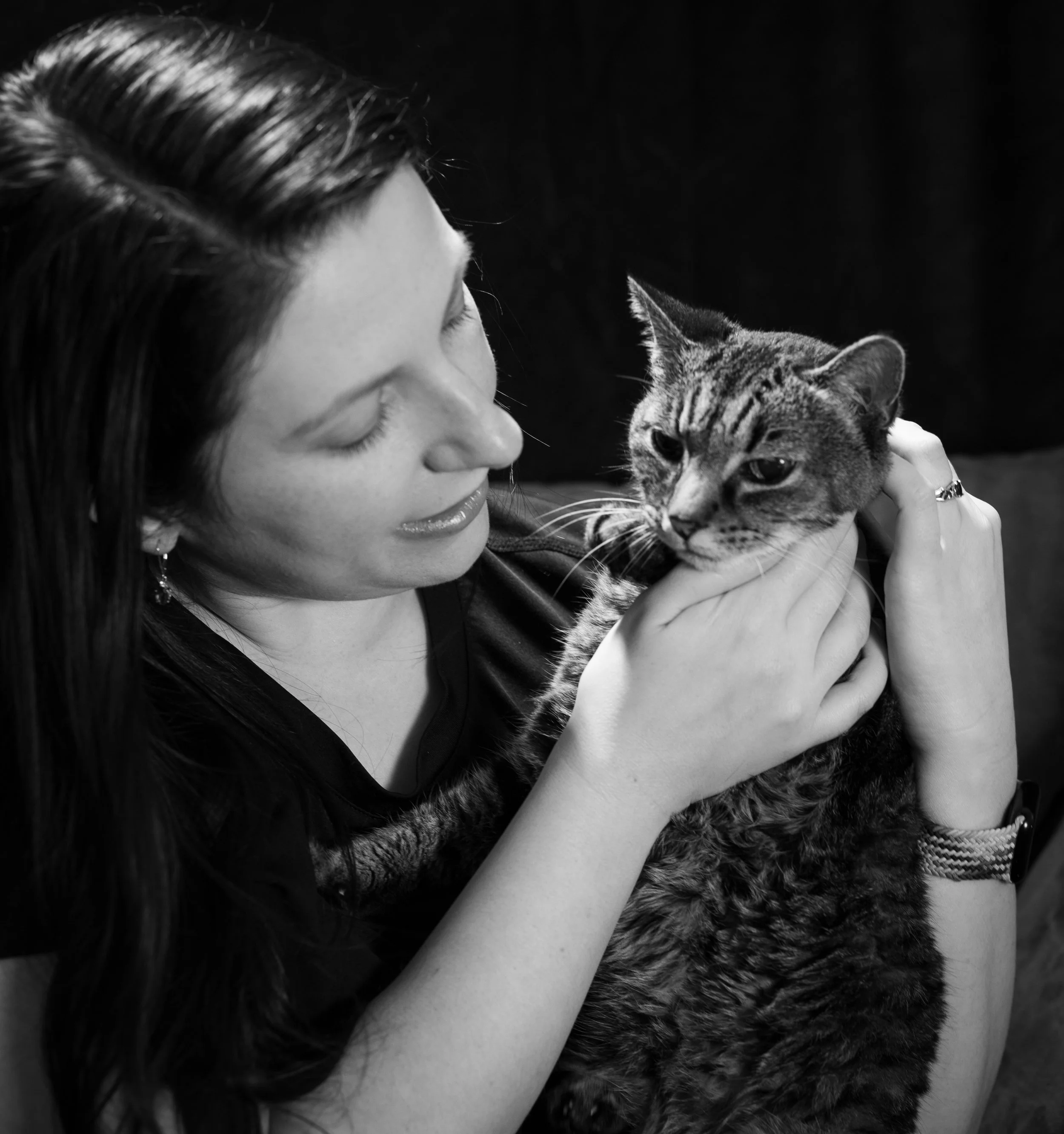 A woman with long dark hair holding a tabby cat close to her face, both looking at each other in an intimate moment.