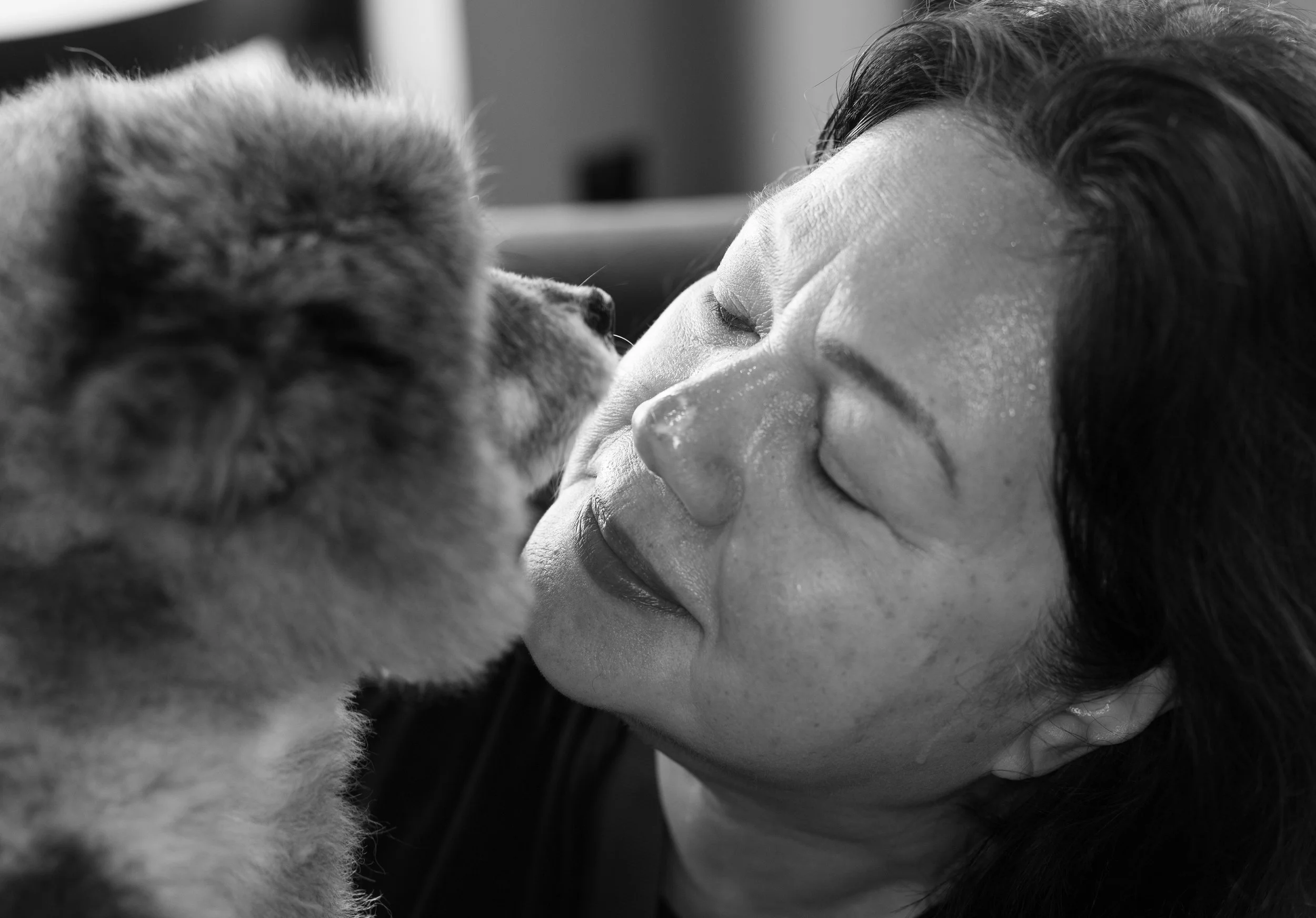 A woman with dark hair, lying close with her eyes closed, faces a dog with fluffy fur, nose touching nose, in a close-up black and white photograph.