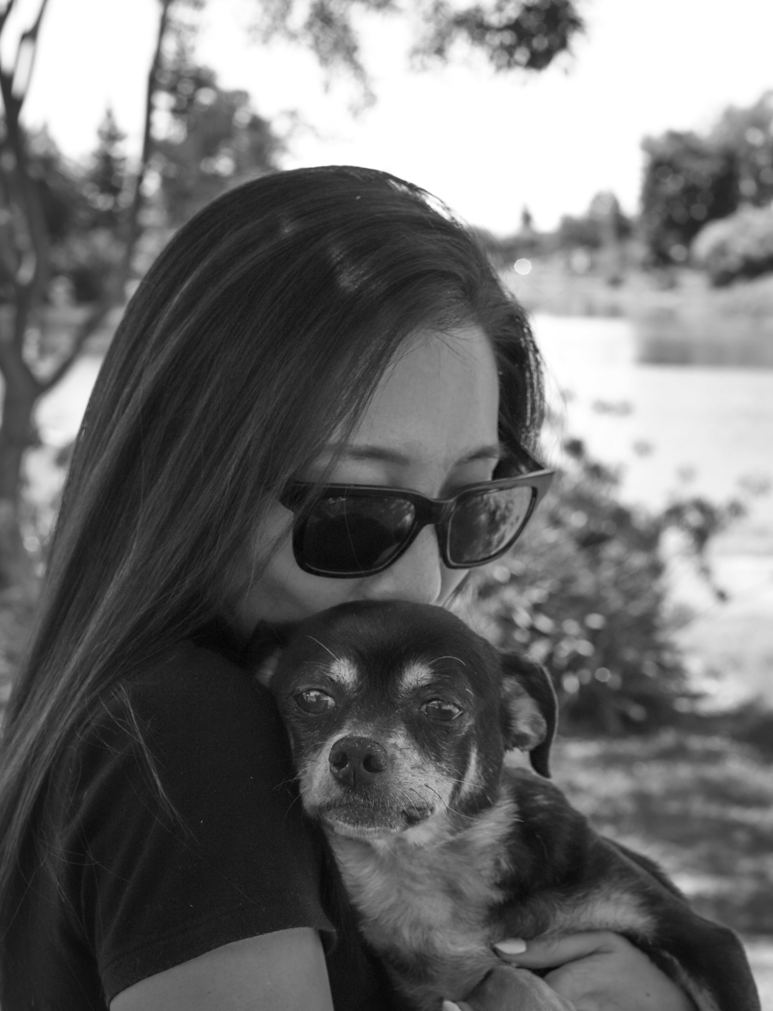 A woman with long hair wearing sunglasses is hugging a small dog outdoors, with trees and a body of water in the background.
