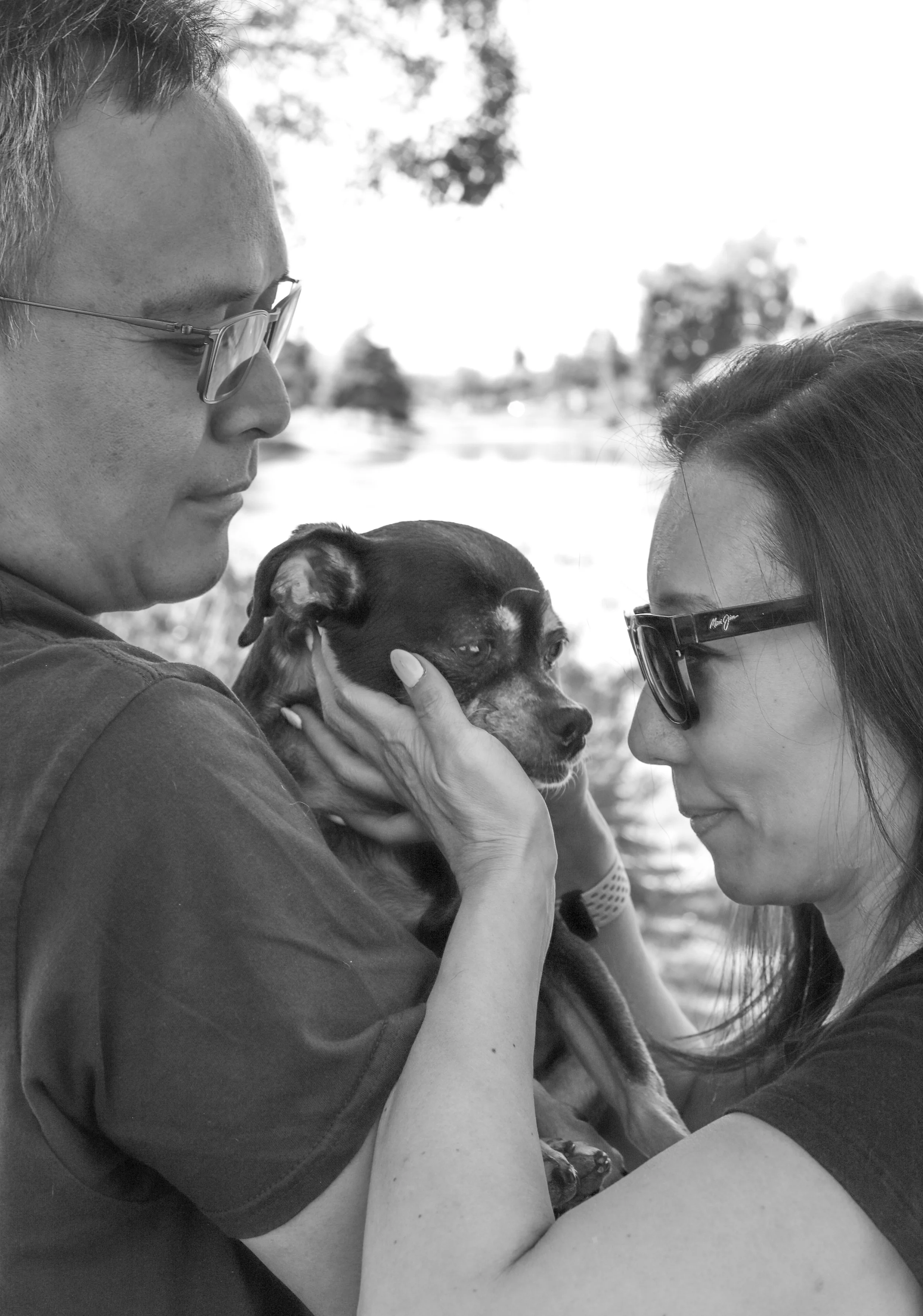 A black and white photo of a man and woman facing each other, holding a small dog close to their faces outdoors. The man is on the left, wearing glasses, and the woman is on the right, also wearing glasses. The dog, which has a dark coat with some li