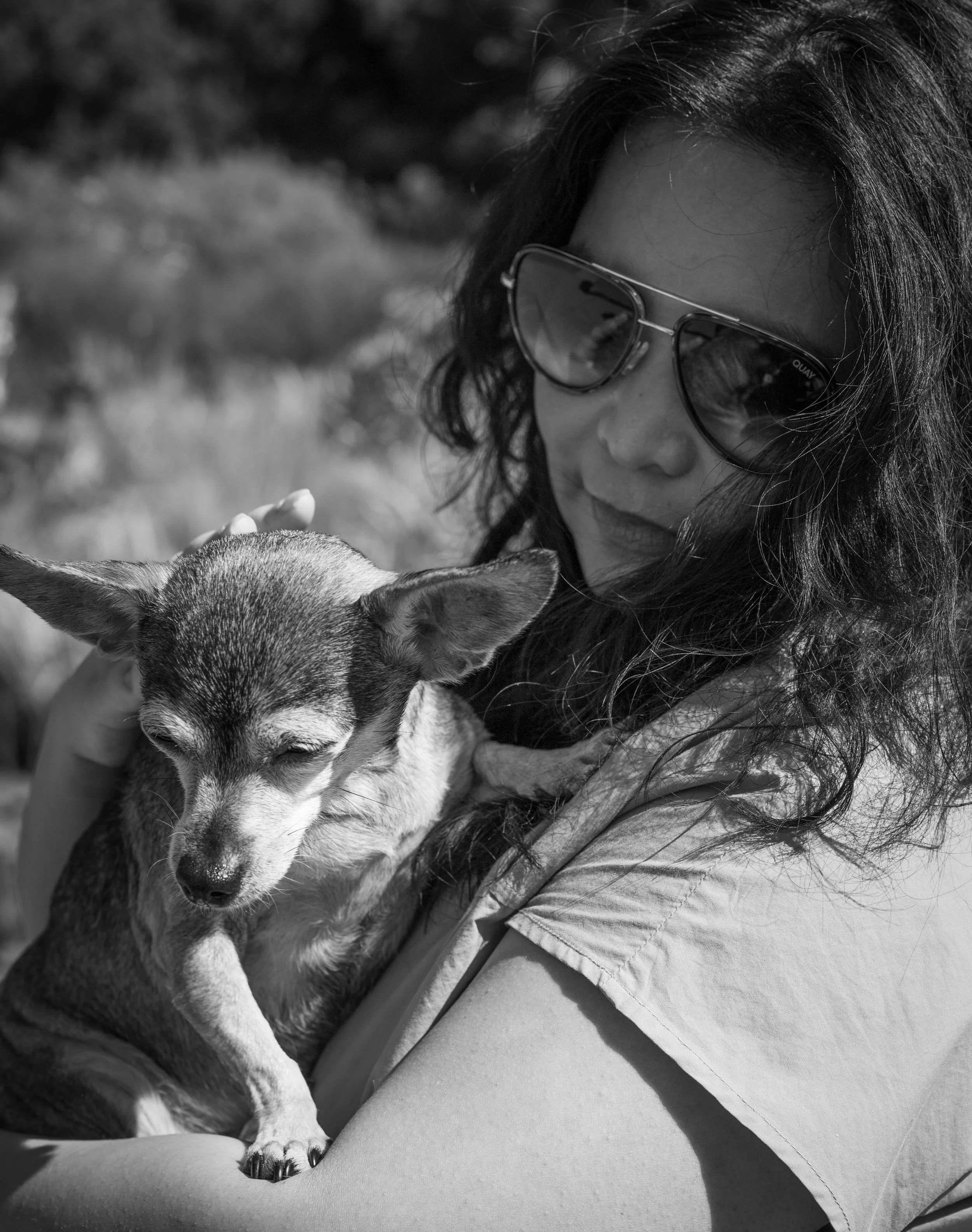 A woman with dark hair and sunglasses holding a small dog with pointed ears in her arms outdoors.