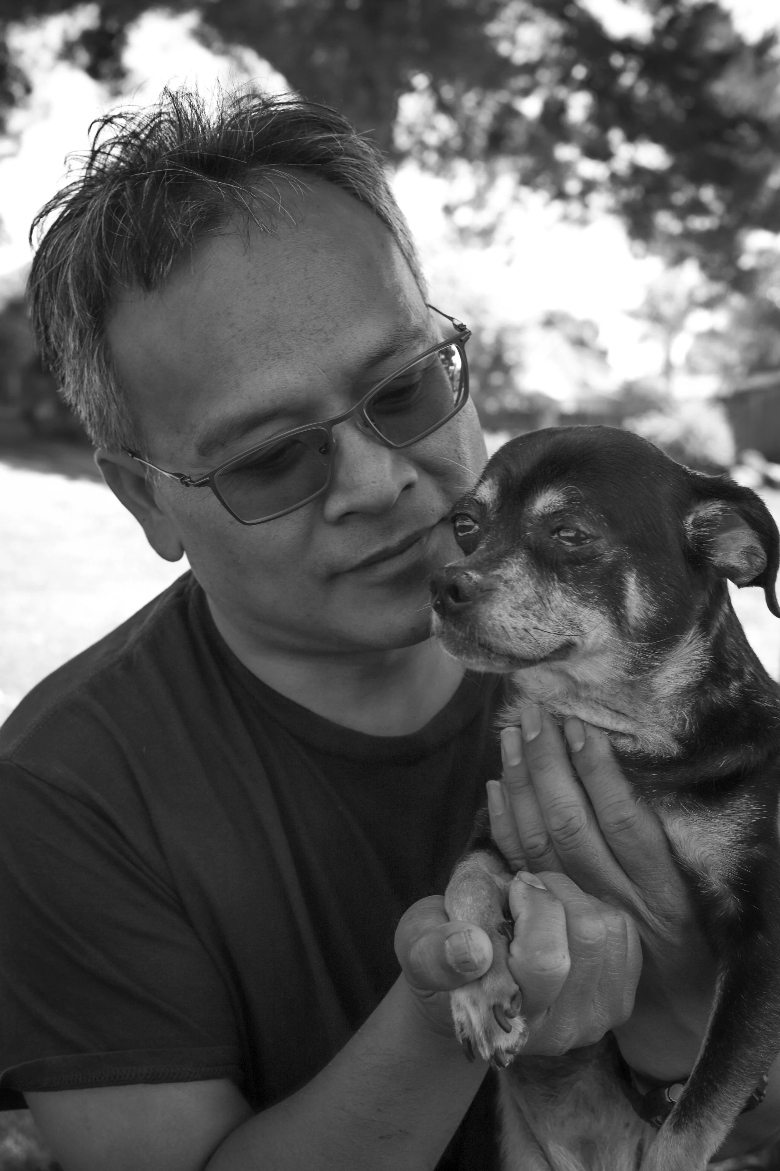A person wearing sunglasses holds a small dog, both appearing content with their faces close together outdoors with trees in the background, in a black-and-white photo.
