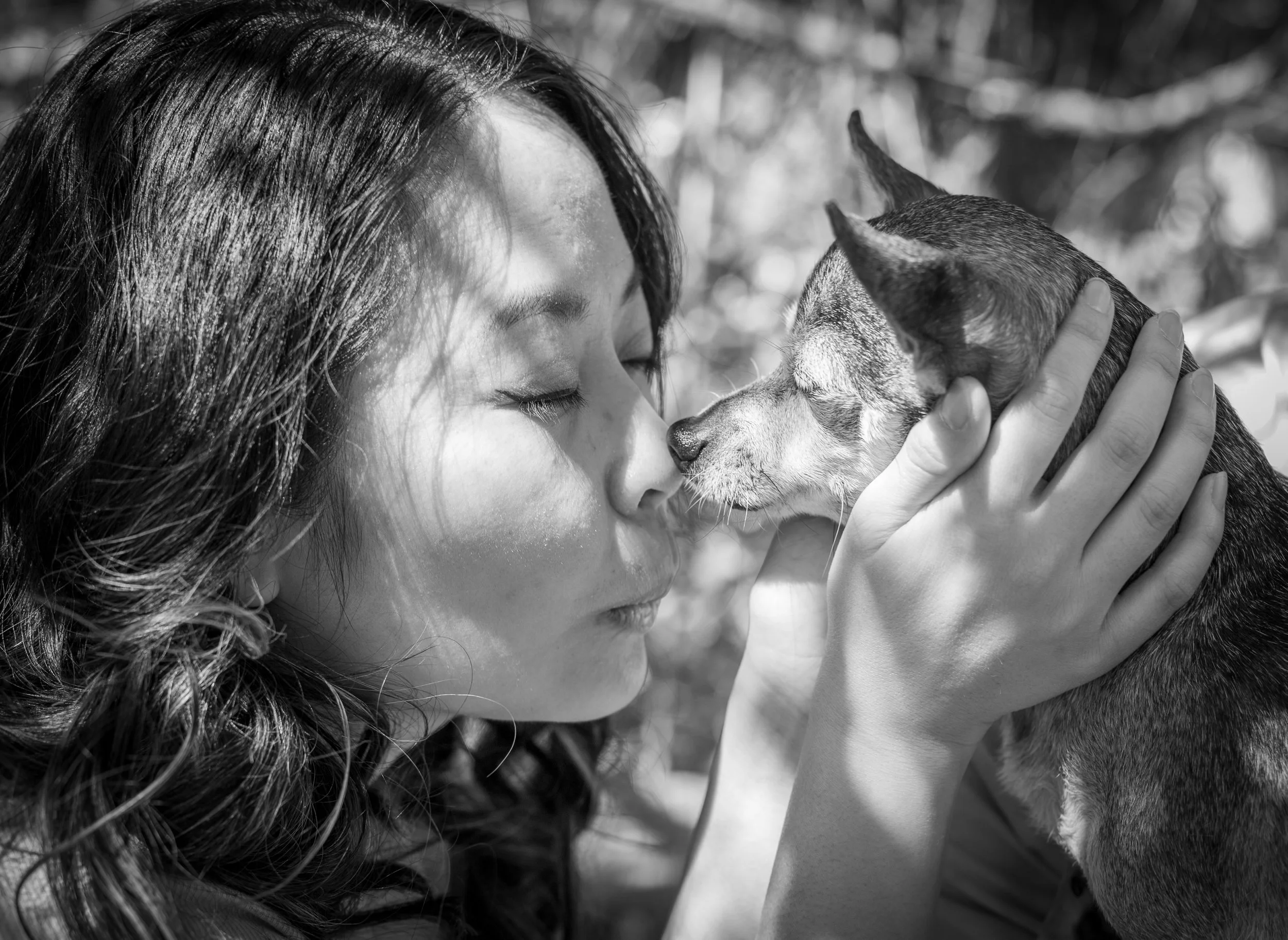 Black and white photo of a woman with long hair gently holding a small dog close to her face, both with eyes closed touching noses.