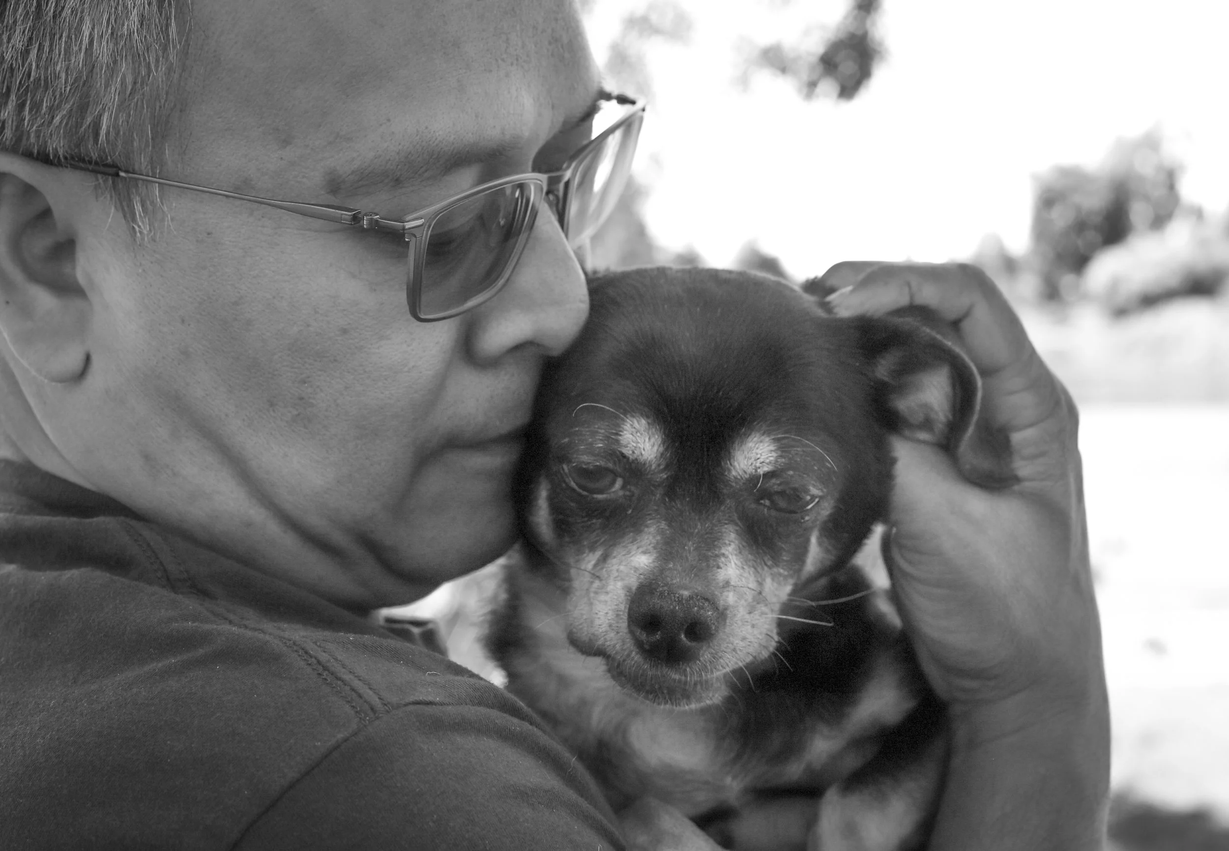 A person wearing glasses holding and gently cuddling a small dog with a sad expression, black and white photo.