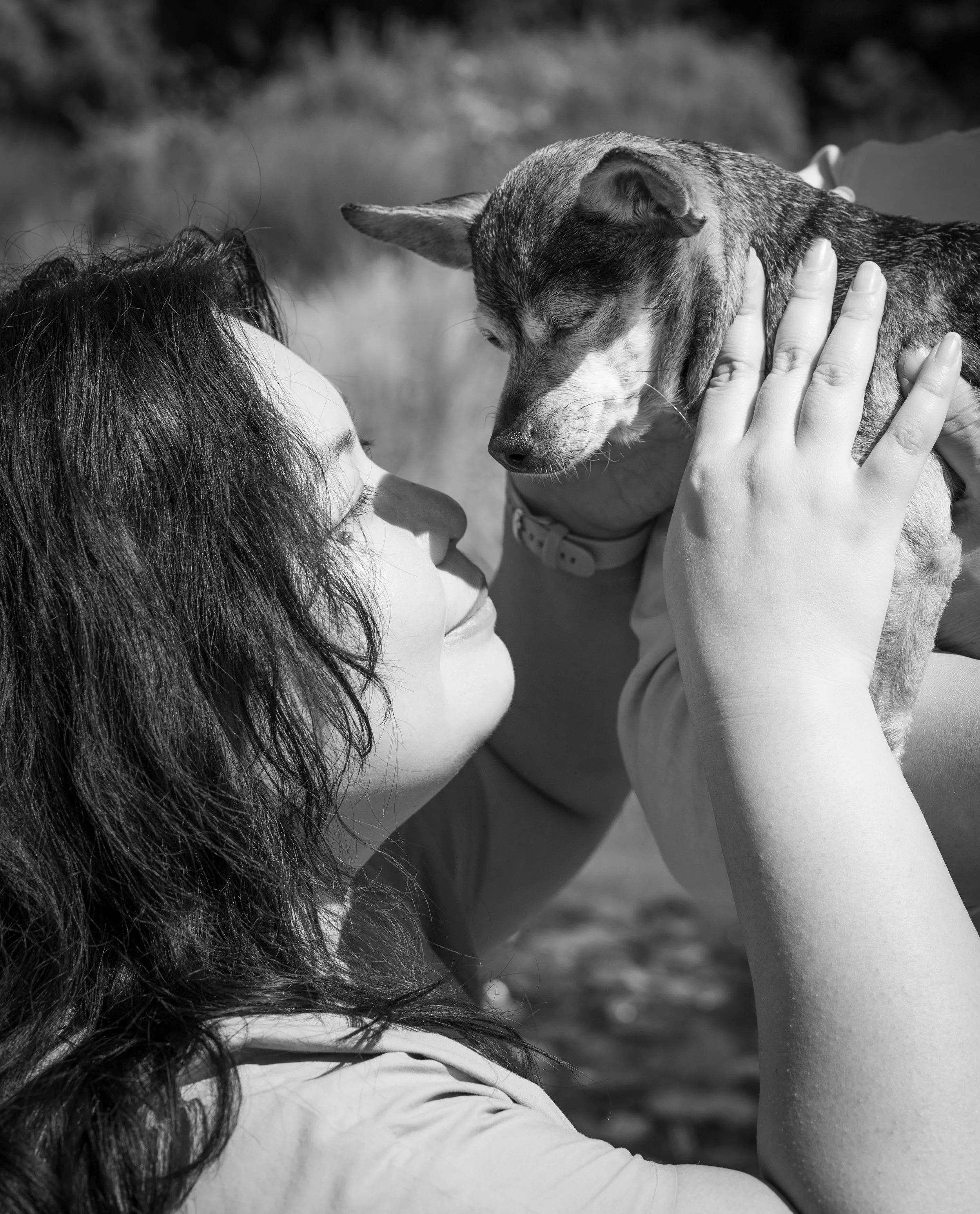 A woman with curly hair holding a small dog close to her face outdoors, both with eyes closed, touching noses in a black and white photo.