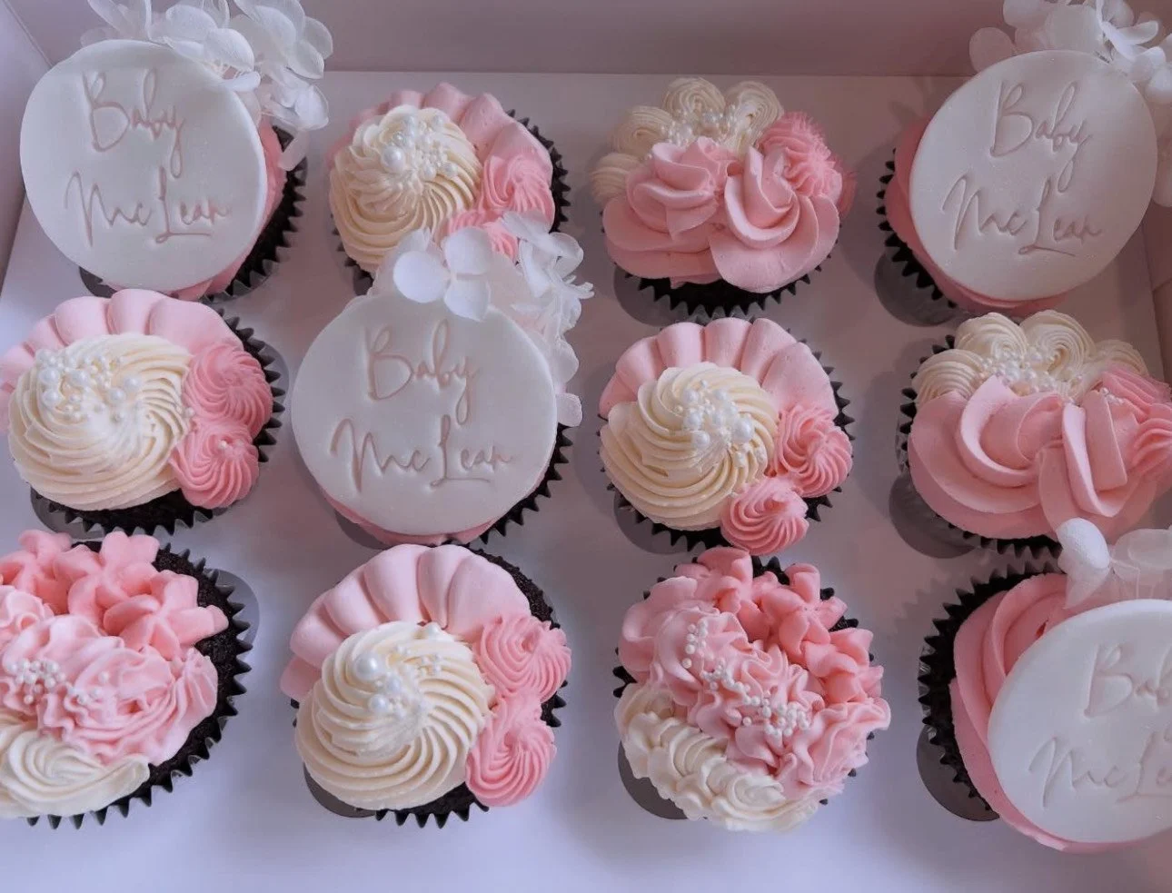 A tray of cupcakes decorated with pink and white frosting, some with white circular toppers that say 'Baby M soleil,' and others with piped flowers and decorative swirls.