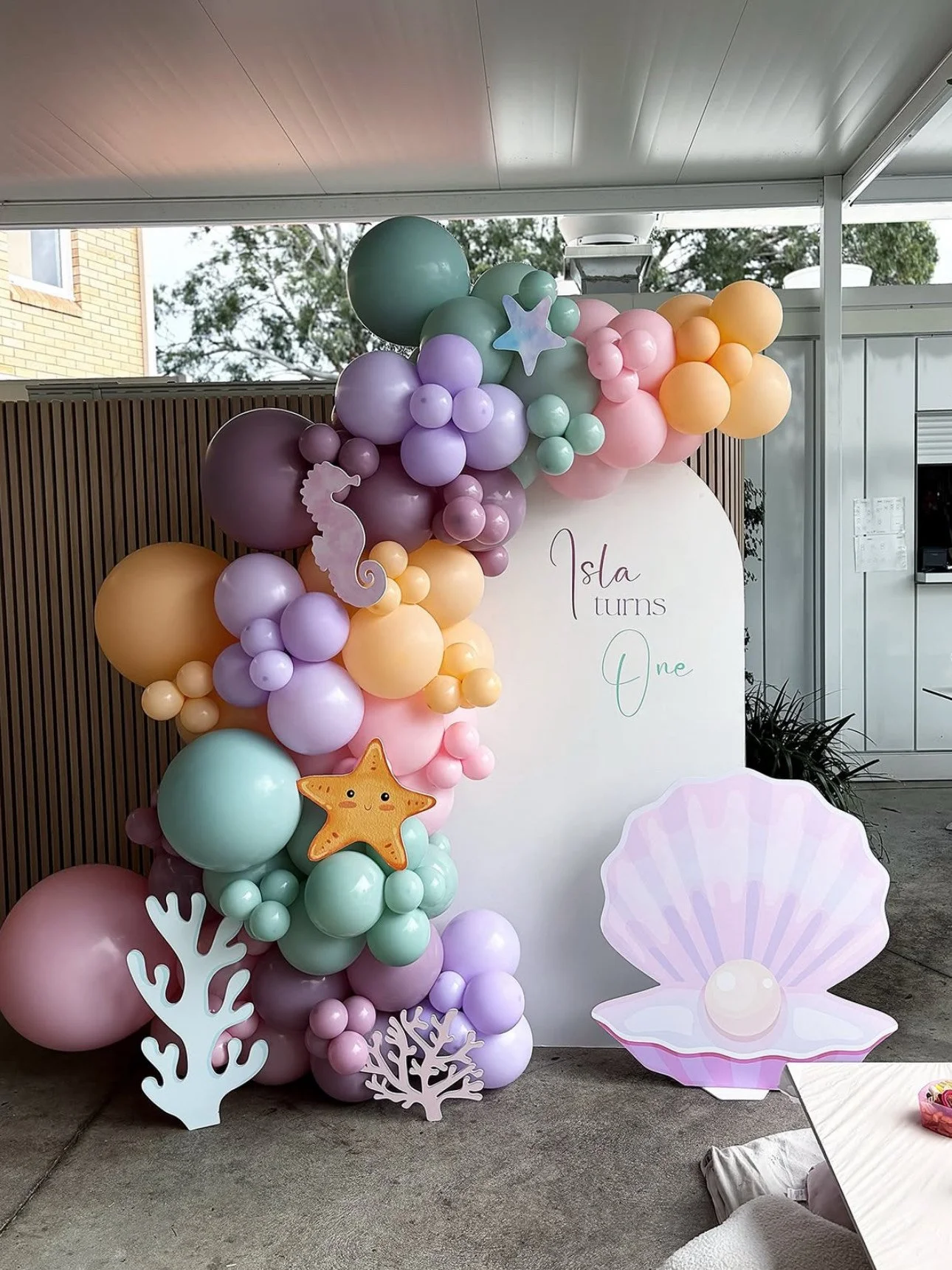 Balloon arch with pastel colors, ocean-themed decorations, and a sign that reads 'Isla turns One' for a first birthday celebration.