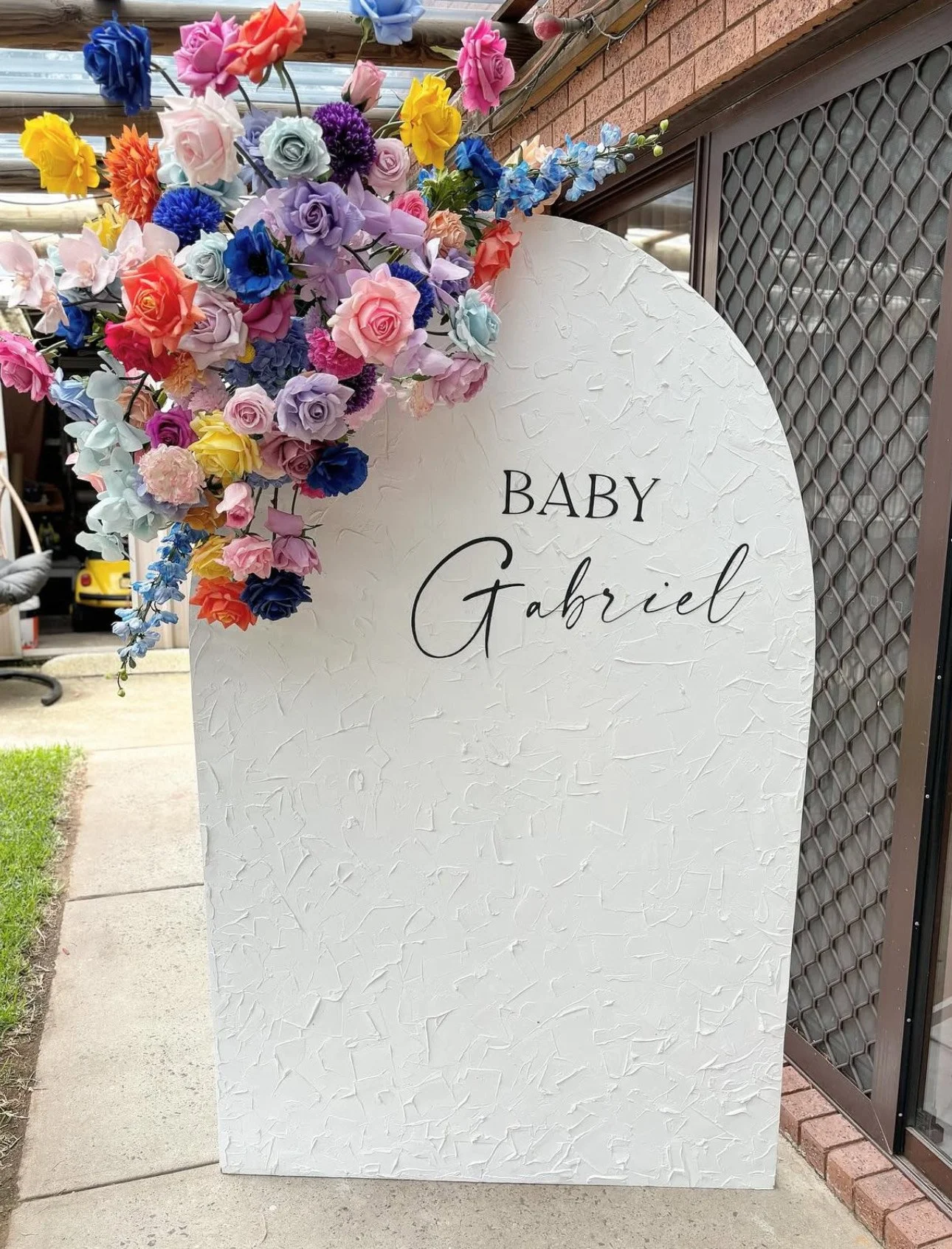 A white textured signboard with black cursive and capital lettering that reads 'BABY Gabriel,' decorated with colorful artificial flowers in pink, purple, blue, yellow, and orange, placed outdoors next to a building and sidewalk.