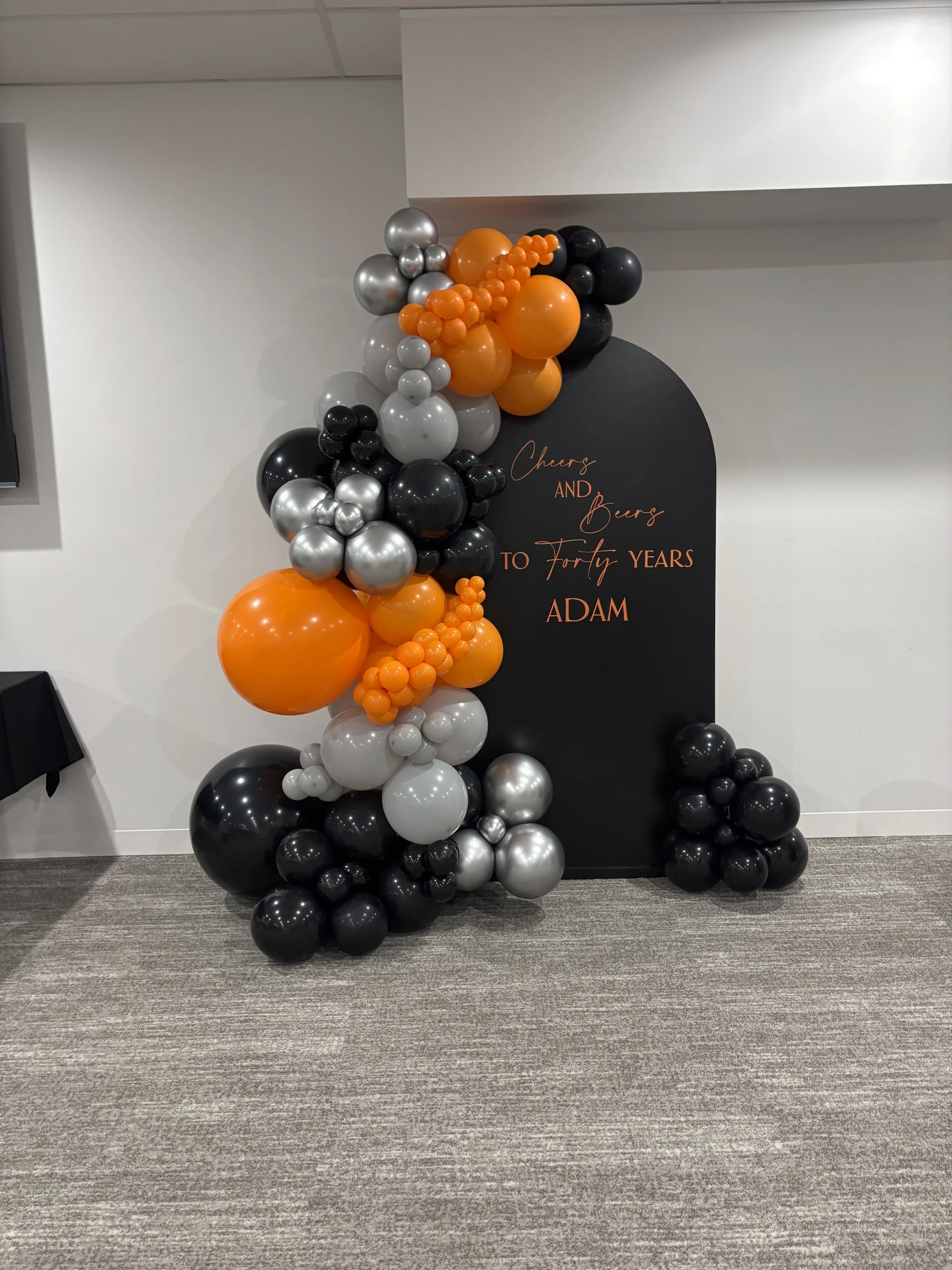Balloon arrangement with black, silver, orange, and gray balloons next to a black backdrop with orange text celebrating a forty-year milestone for Adam.