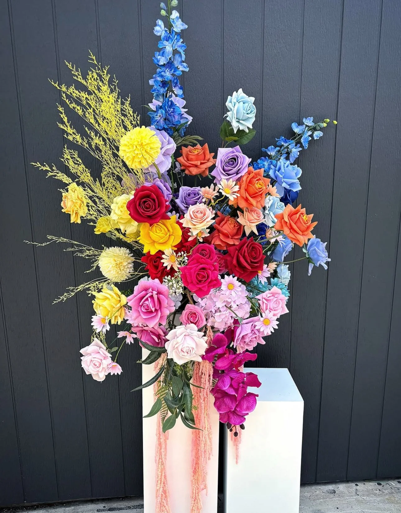 Colorful flower arrangement with roses, orchids, and other flowers in a pink and white vase on a white pedestal against a dark wall.