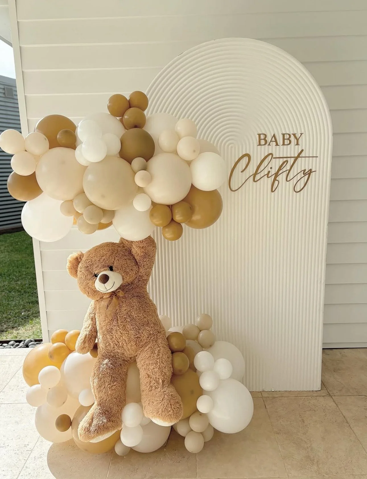 Decorated baby shower setup with a teddy bear holding a cluster of beige, white, and gold balloons in front of a white panel with the text 'Baby Clifty'.
