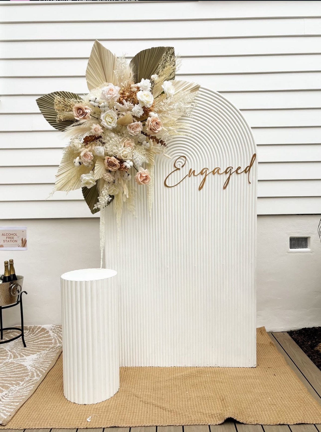 A wedding or engagement display with a white backdrop that says "Engaged" in gold lettering, decorated with a floral arrangement of cream, blush, and white flowers, and dried leaves, mounted on the top left side of the backdrop. There is a small white pedestal in front of the backdrop with a textured, ribbed design. To the left, a table with beverages and a sign indicating an alcohol-free station is visible.