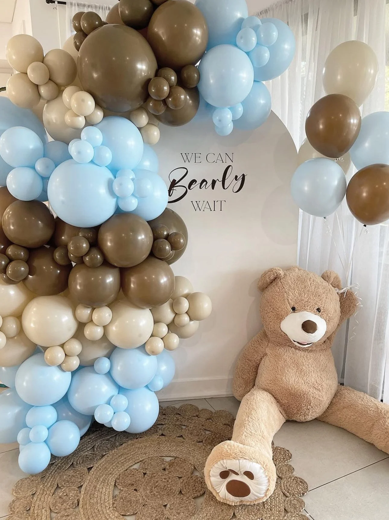 Decorative display for a baby shower or gender reveal with blue, ivory, and brown balloons, a large teddy bear, and a sign that says "We can barely wait."