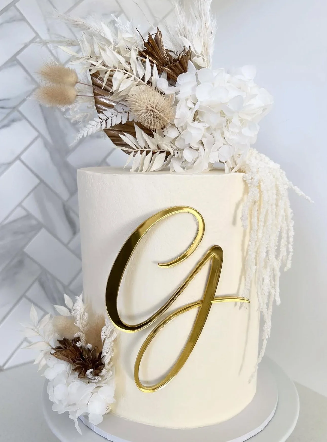Cream-colored cake decorated with white and beige dried flowers, featuring a gold cursive initial 'G' on the front.