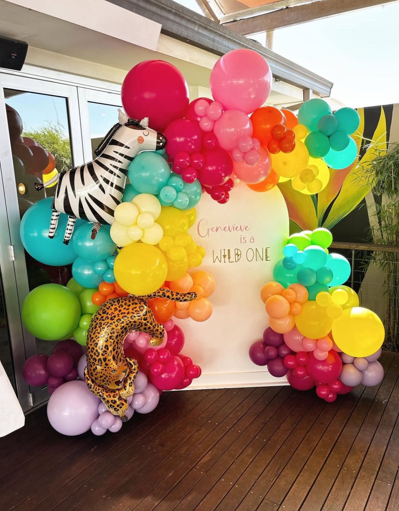 Colorful balloon display with a zebra and a cheetah balloon, featuring a white backdrop with the phrase "Genevieve is a WILD ONE" written on it.