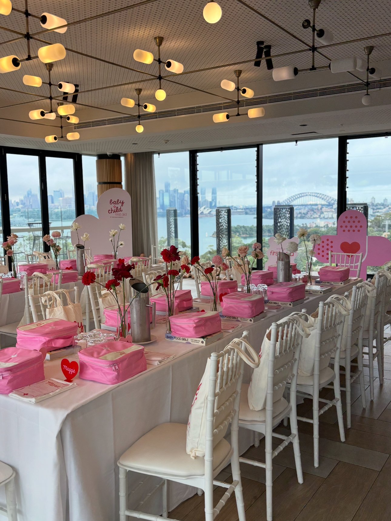 Decorated indoor event space with tables covered in white tablecloths, set with pink boxes, pink and red flowers, and baby-themed signs, with large windows showing a city skyline and water outside.
