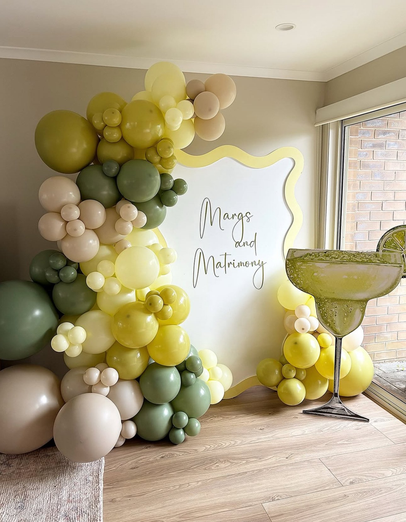 Decorative display with green, beige, and cream balloons arranged around a white backdrop with yellow wavy frame and the words 'Margs and Matrimony' written on it, next to a large margarita glass with a lime garnish.