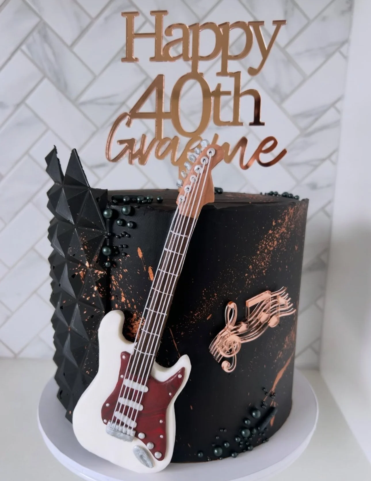 A black birthday cake with music-themed decorations, including a white and red electric guitar, black geometric shapes, black pearls, a rose gold musical note, and a "Happy 40th Gorgeous" topper.
