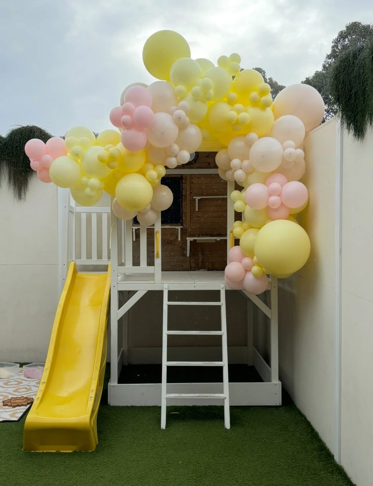 Children's playhouse decorated with yellow, pink, and white balloons, and a yellow slide.