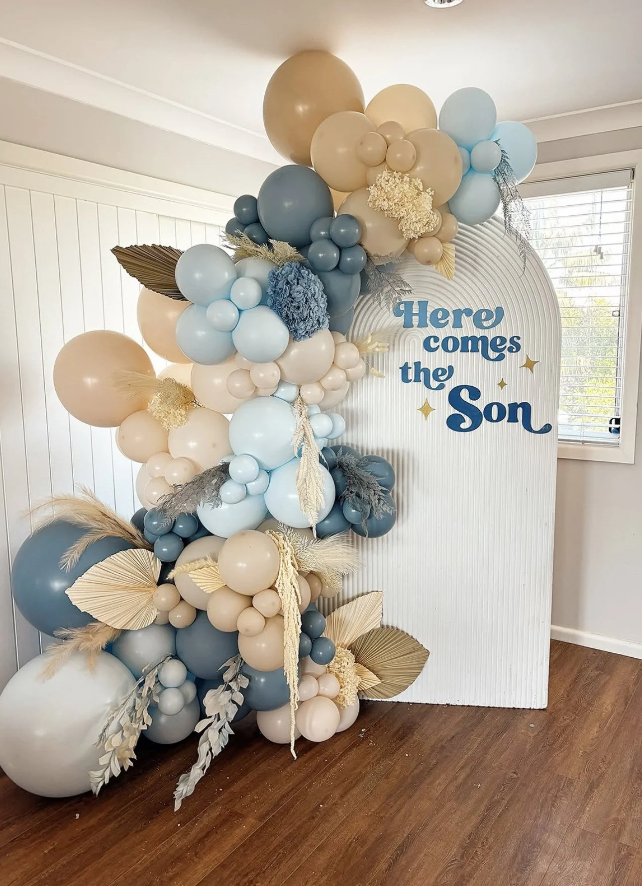 Decorative balloon arrangement in shades of beige, blue, and white with gold and beige accents, featuring various sizes of balloons, artificial leaves, dried flowers, and pampas grass, with a white sign reading "Here comes the Son" in blue letters, set against a white panel background near a window with blinds.