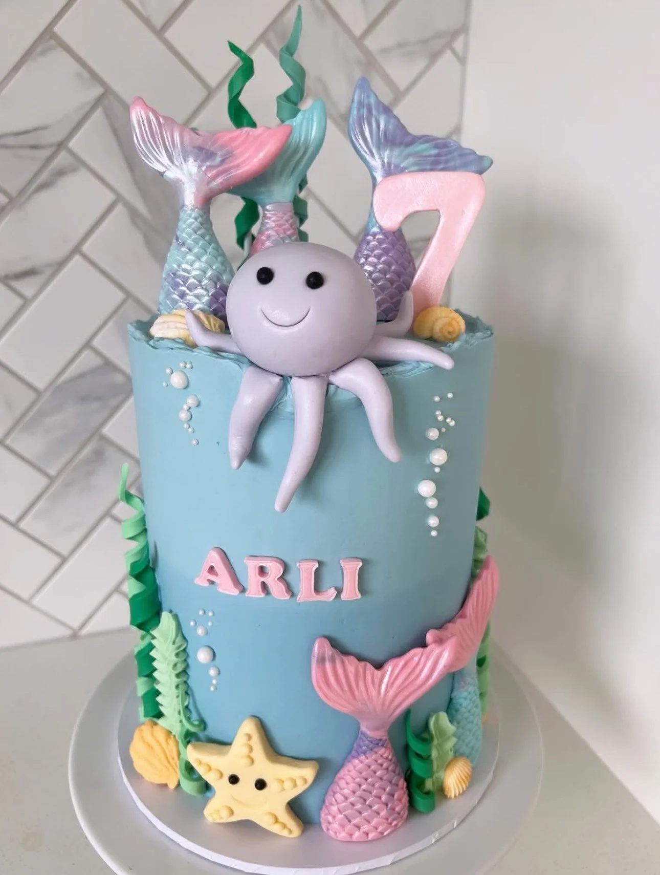 A mermaid-themed birthday cake decorated with pastel-colored mermaid tails, a smiling octopus topper, and seashells. The cake has the name 'Arli' written on it.