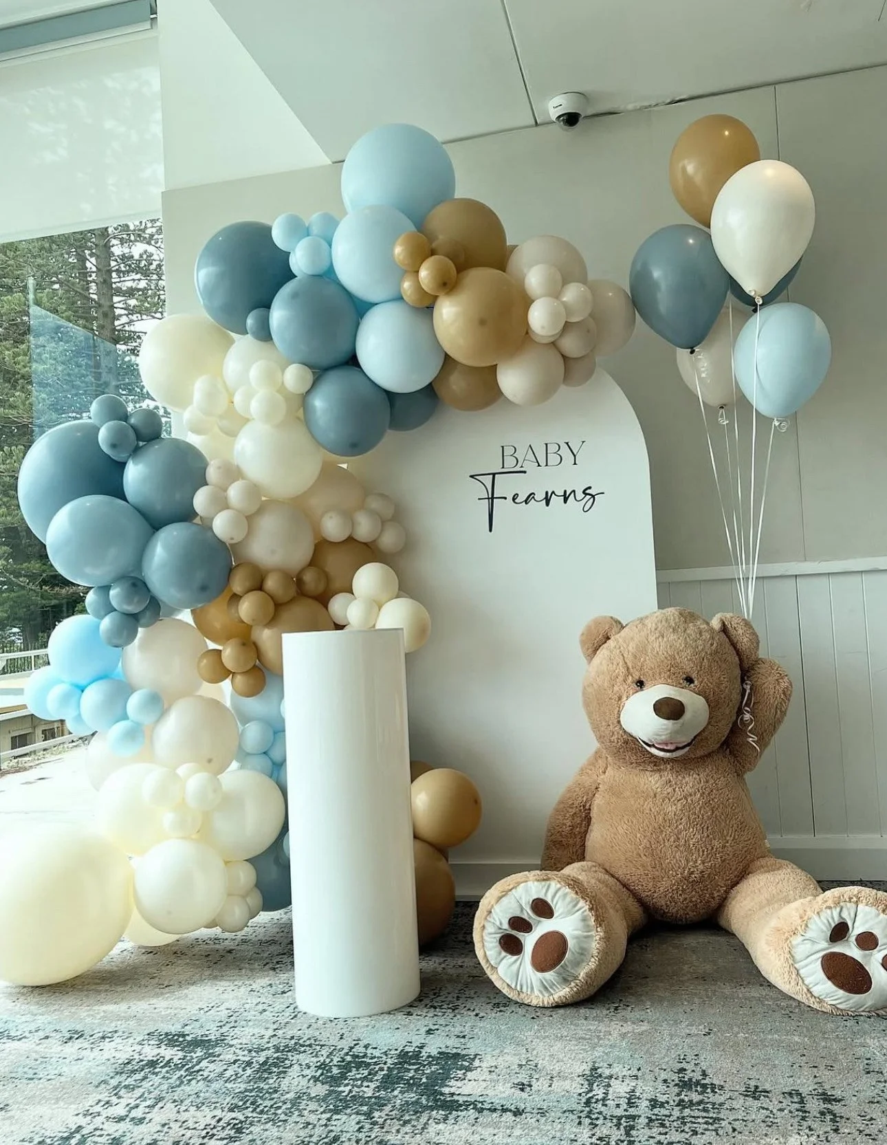 Decor for a baby shower with a large teddy bear, balloons in shades of blue, white, and tan, and a sign that reads 'Baby Fears' in a white arch background.