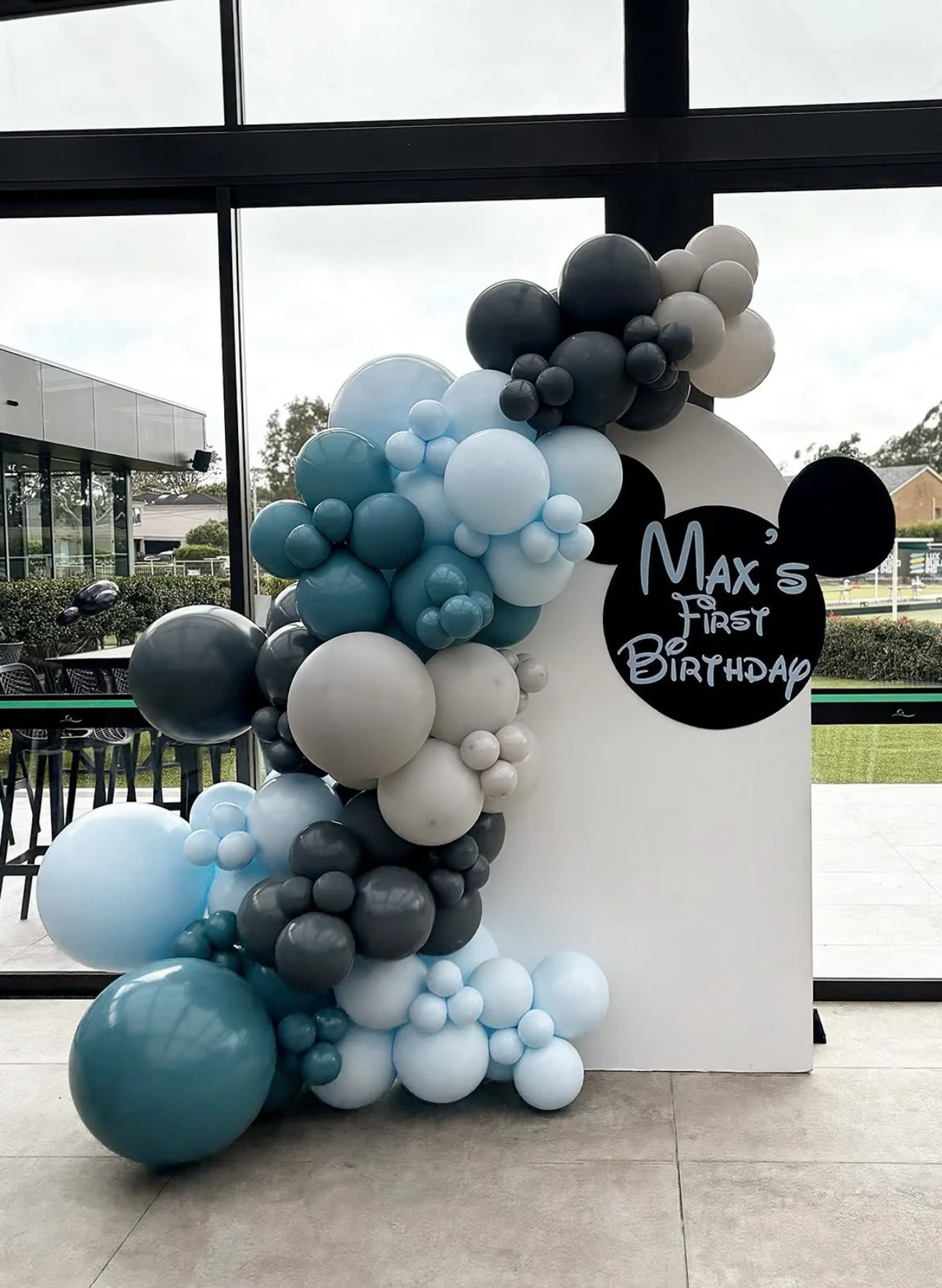 Balloon arrangement with black, white, and blue balloons next to a white stand with a sign that reads "Max 5 First Birthday," decorated with Mickey Mouse ears.