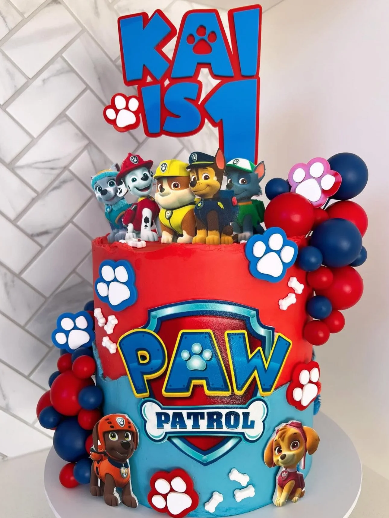 A themed birthday cake for a child's first birthday featuring characters from Paw Patrol, decorated with paw prints, balloons, and a toppper reading 'Kai is 1'.