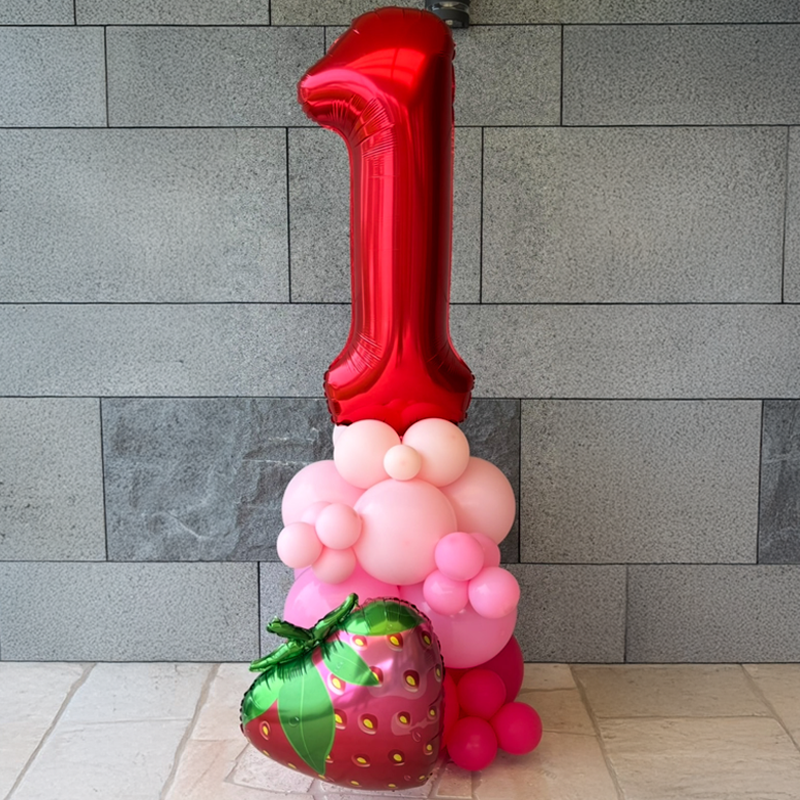 A balloon arrangement featuring a large red number '1' balloon at the top, pink and white balloons below, and a large, decorated strawberry-shaped balloon at the base.
