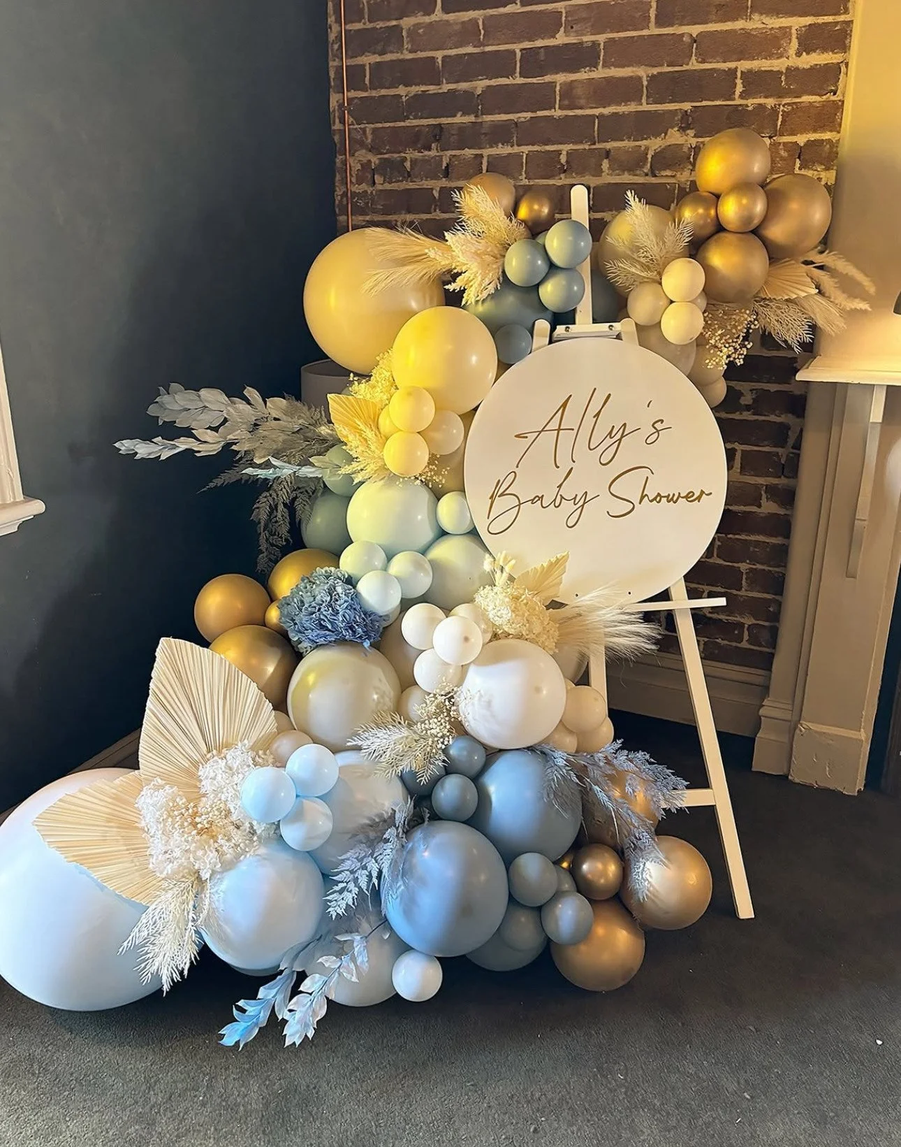 Decorative balloon arrangement in shades of white, cream, gold, and pastel blue with dried leaves and flowers, next to a sign that reads 'Ally's Baby Shower' on an easel, against a brick wall.