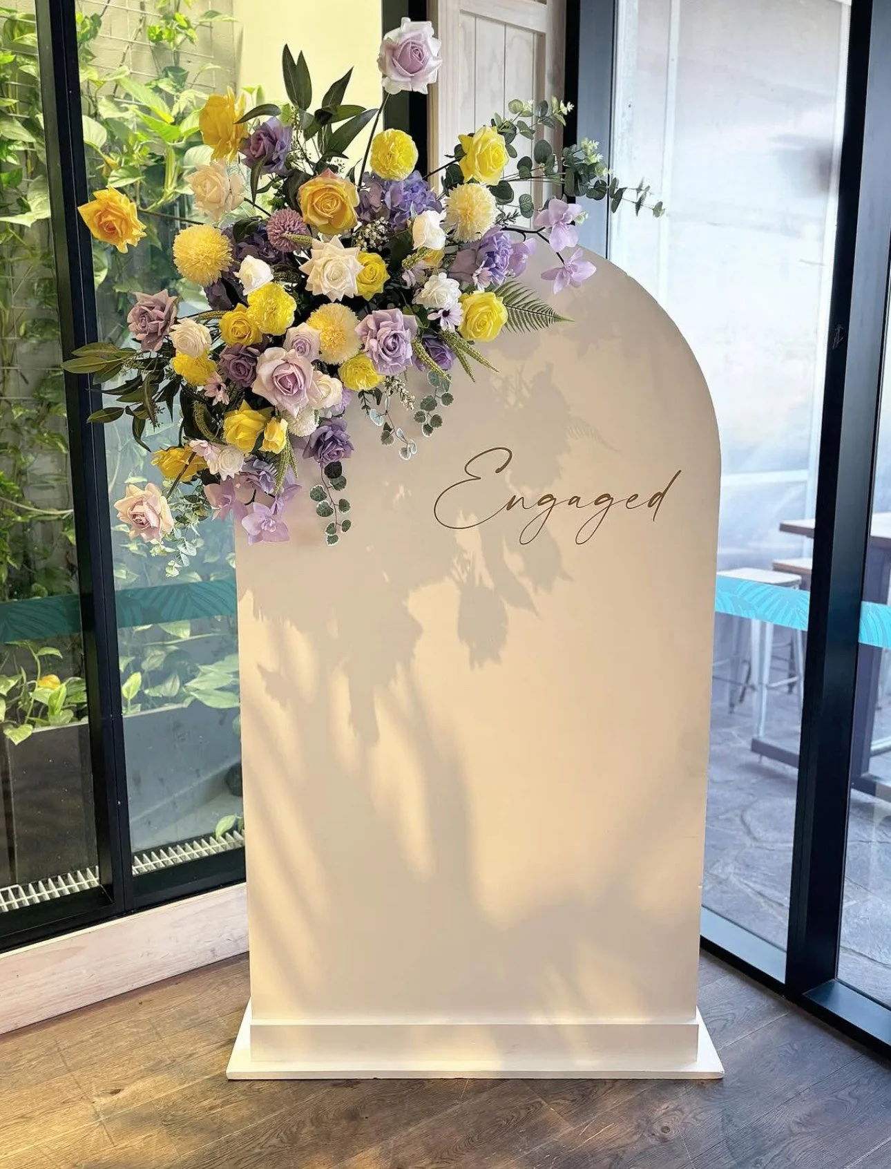 A sign with the word 'Engaged' written on it, decorated with a floral arrangement of yellow, purple, white roses and other flowers, standing on a wooden floor near glass doors.