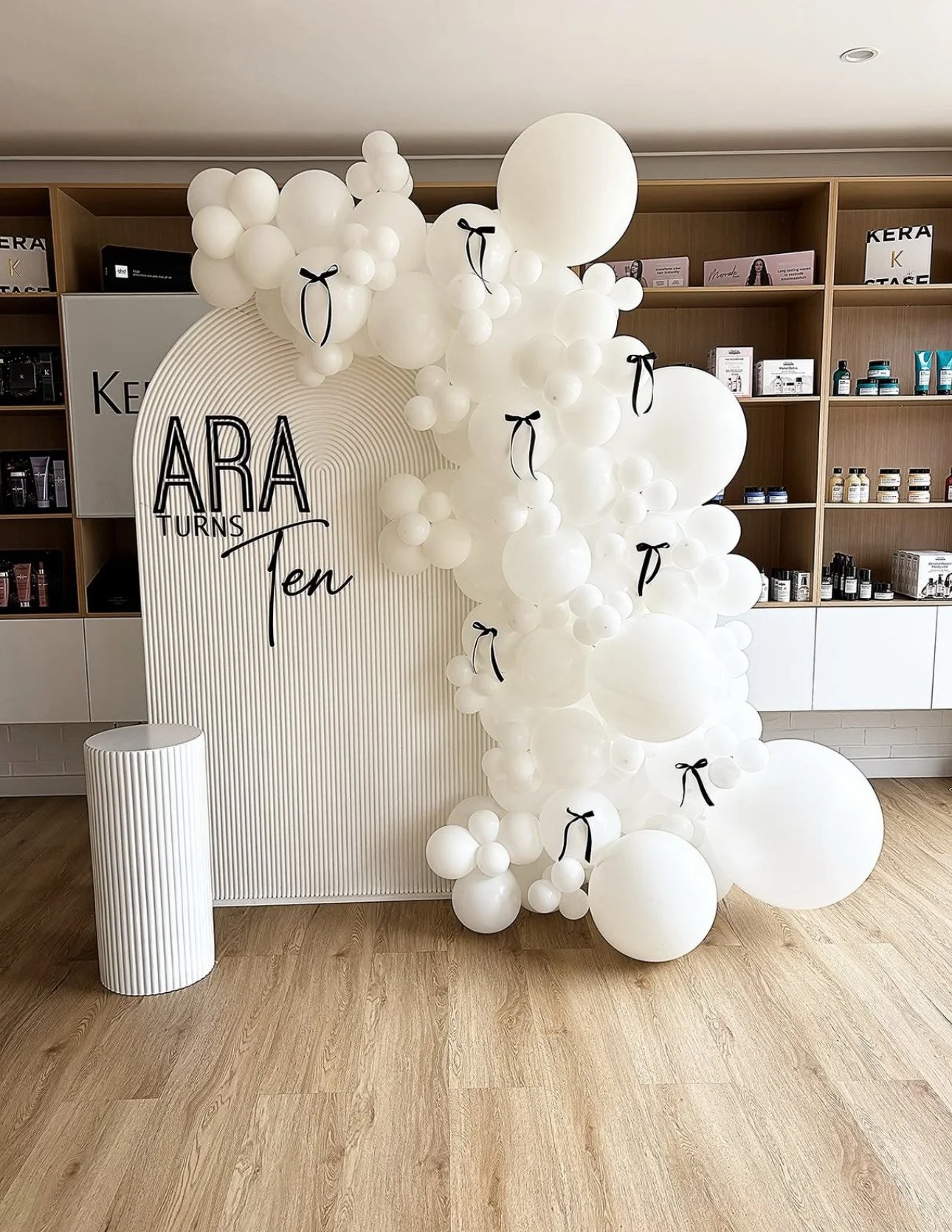 Decorative display celebrating Aria's tenth birthday with white balloons and a backdrop with text 'ARIA TURNS Ten' in front of wooden shelving with products.