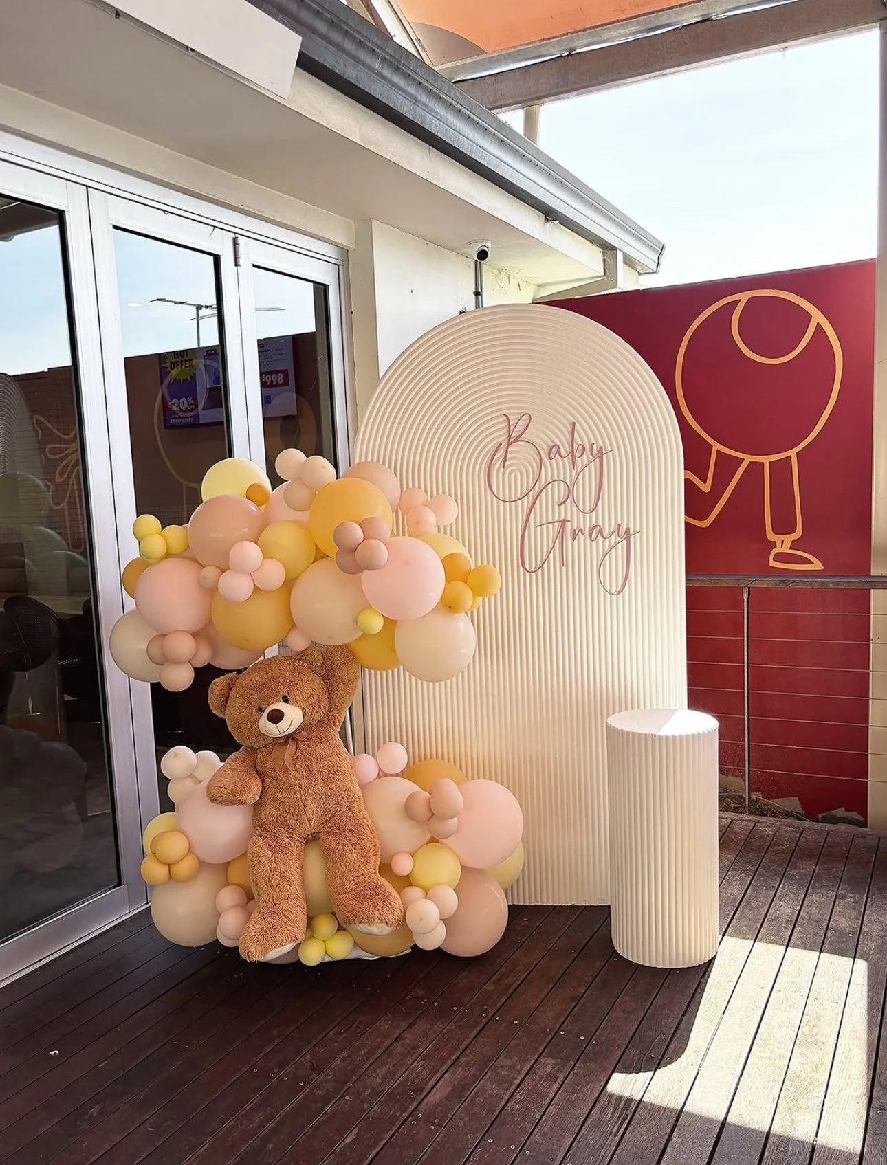 Decorative display with a plush teddy bear sitting among pastel-colored balloons and a sign reading 'Baby Grey' on a white, textured backdrop, set outdoors on a wooden deck.