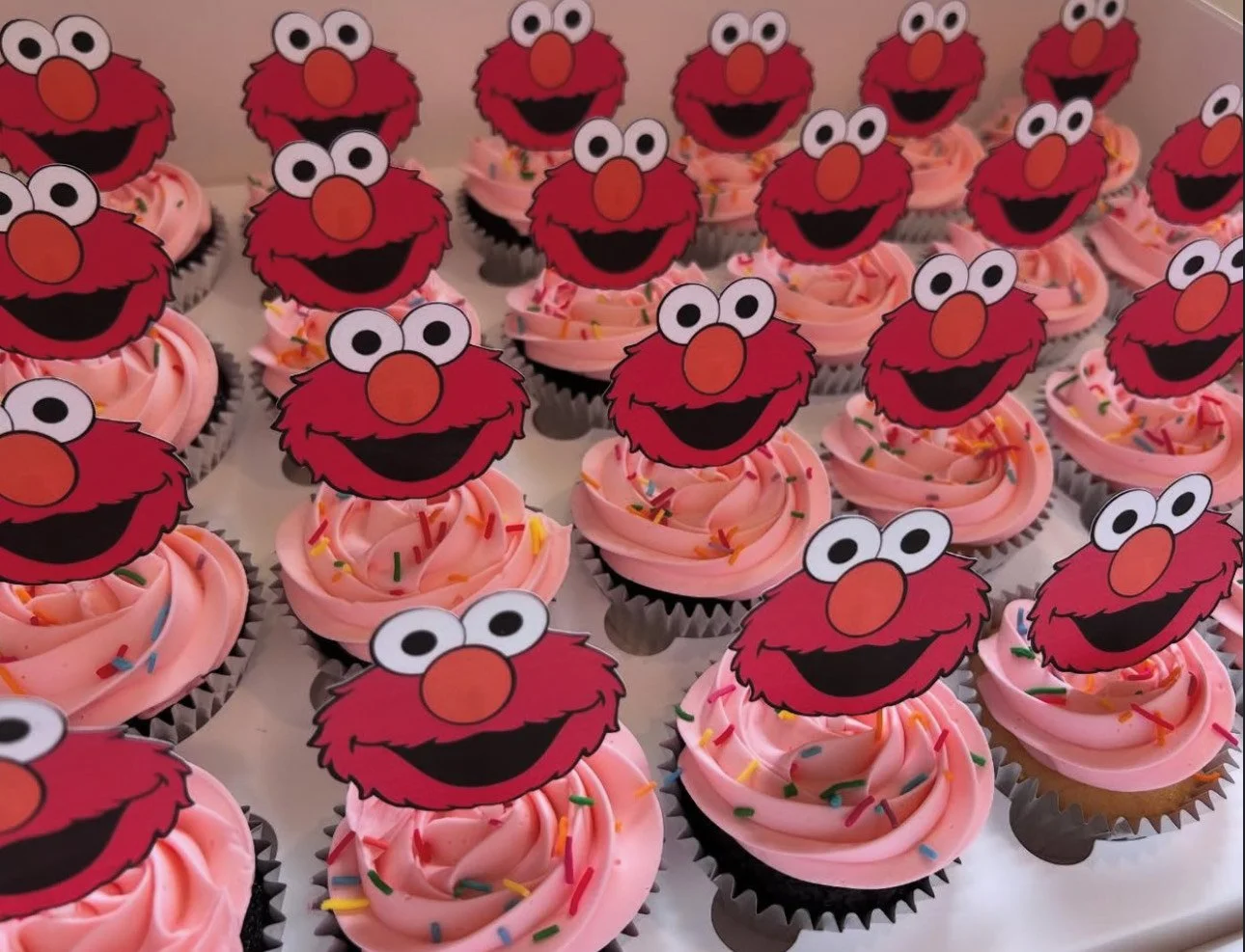 Cupcakes with pink frosting and Elmo themed toppers featuring Elmo's face with large eyes, a red fuzzy face, and an orange nose, topped with multicolored sprinkles.