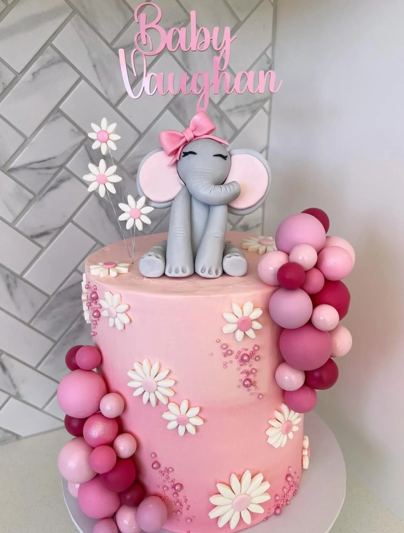 A pink, multi-layered birthday cake decorated with white daisies, pink and red fondant balls, and a gray elephant cake topper with large pink ears, a pink bow, and closed eyes. Pink lettering on top reads "Baby Vaughan."