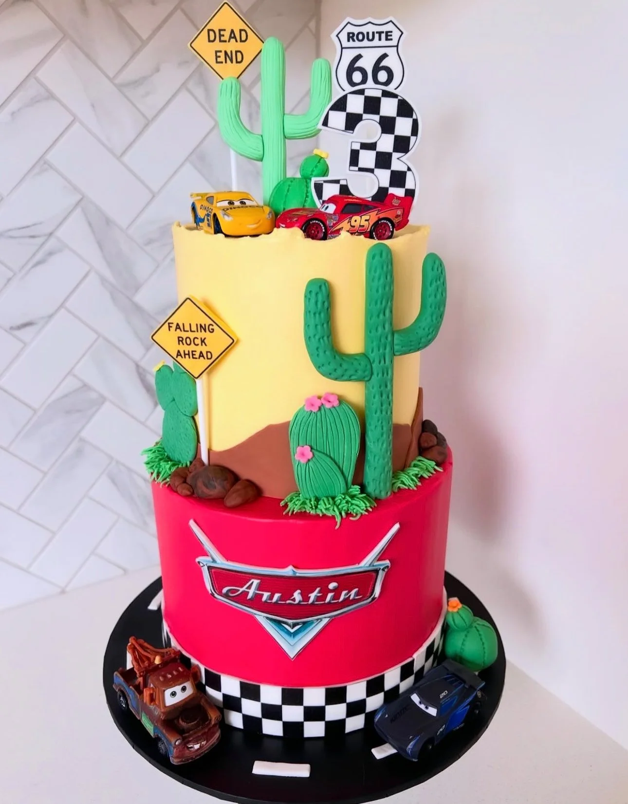A colorful birthday cake with a Cars movie theme, featuring toy cars, cactus decorations, road signs, and an Austin badge, set on a black and white checkered base.