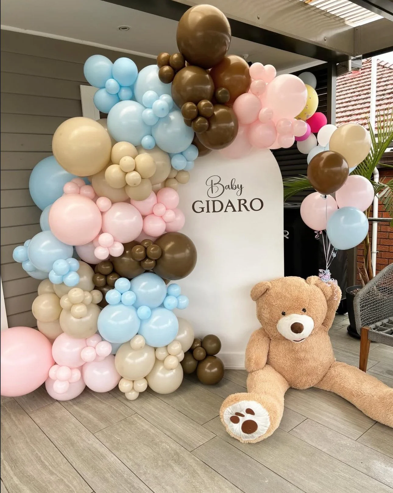 Decorative balloon arch with pastel pink, light blue, beige, and brown balloons surrounding a white backdrop with the text "Baby GIDARO" and a large brown teddy bear sitting on the floor nearby.
