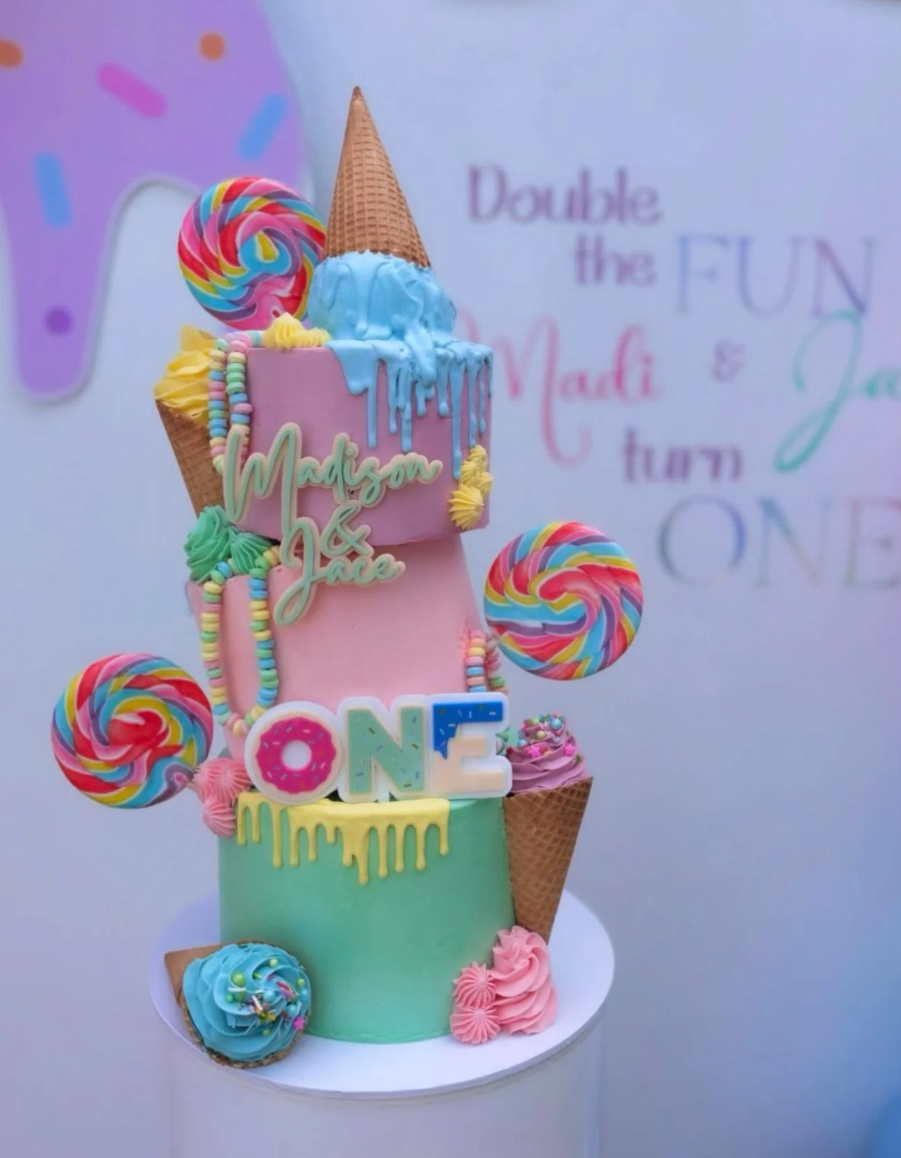 Colorful birthday cake with pastel colors, candy decorations, and an ice cream cone, with the words "Madison & Jace" and "ONE" on it.
