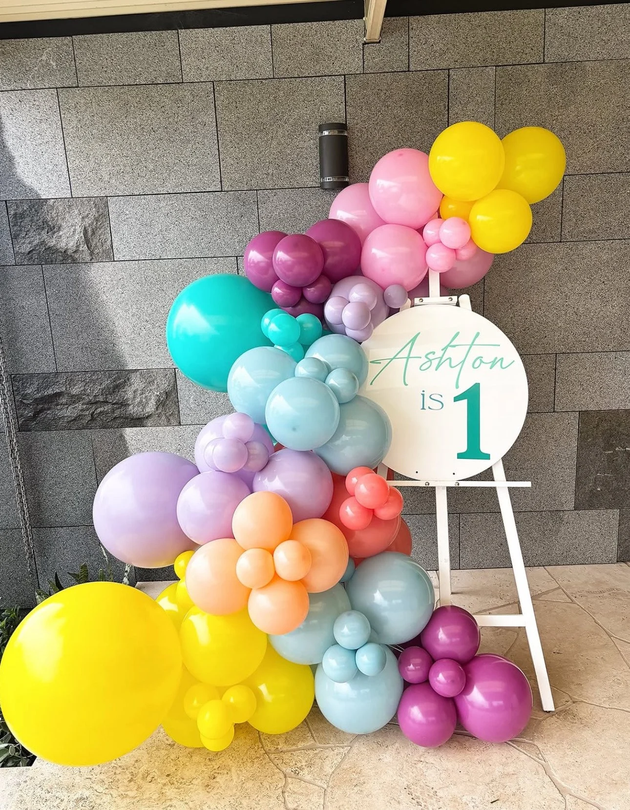 Colorful balloon arrangement with a sign that says "Ashton is 1" for a first birthday celebration.