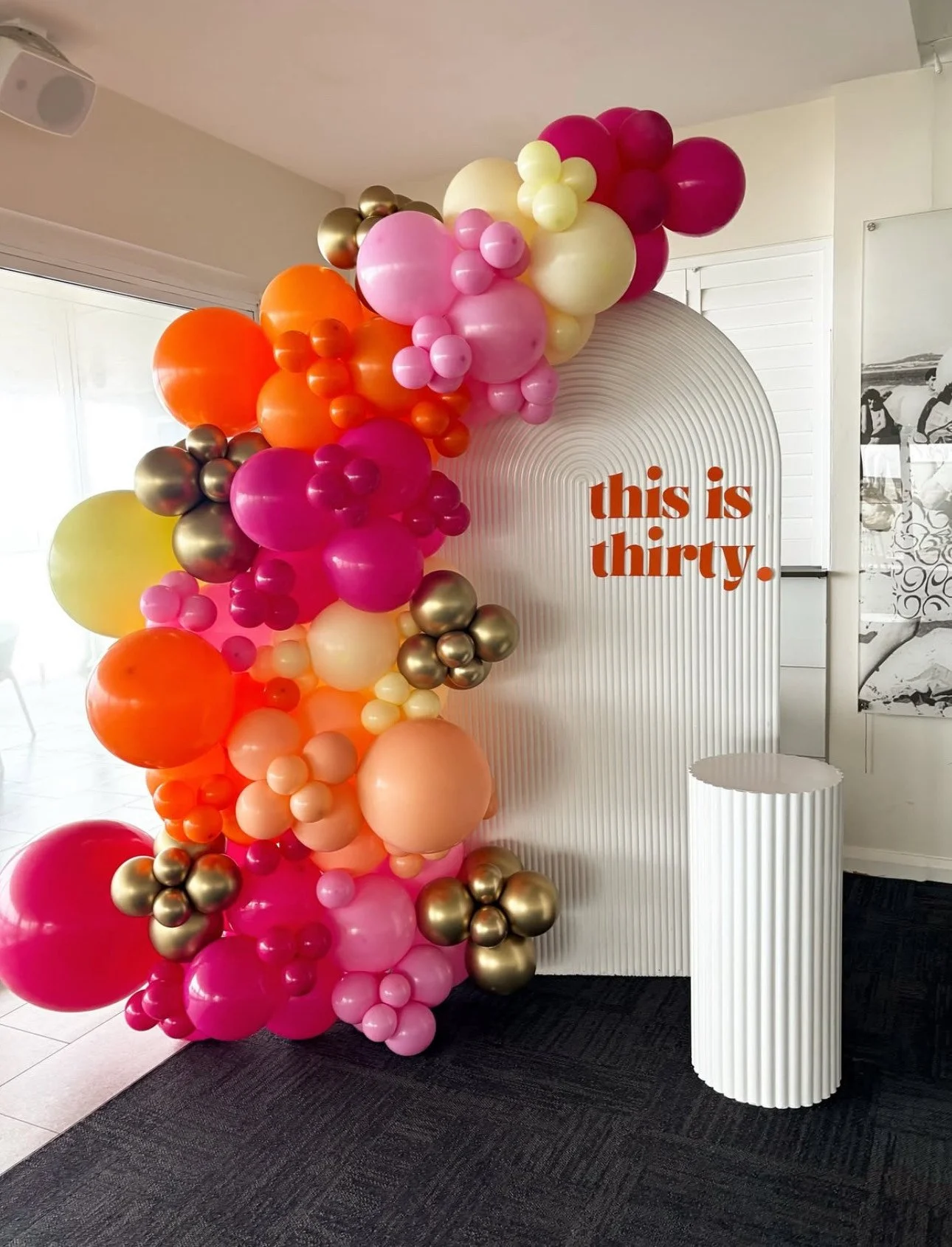 Colorful balloon arrangement in shades of pink, orange, yellow, and gold next to a white, ribbed backdrop with the orange text 'this is thirty.' on it.