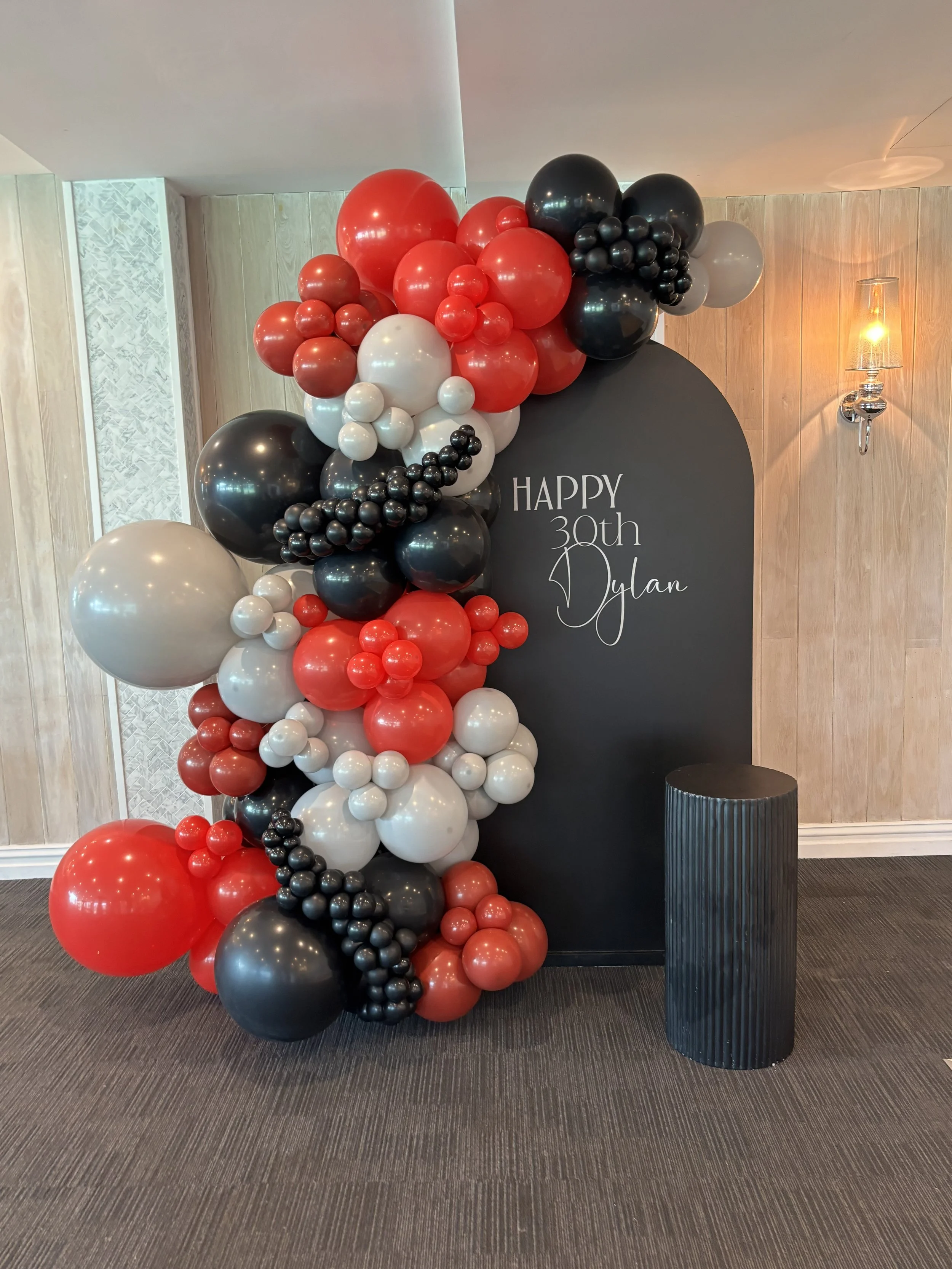Balloon arch with red, black, and white balloons next to a black backdrop with white text that reads 'Happy 30th Dylan'.