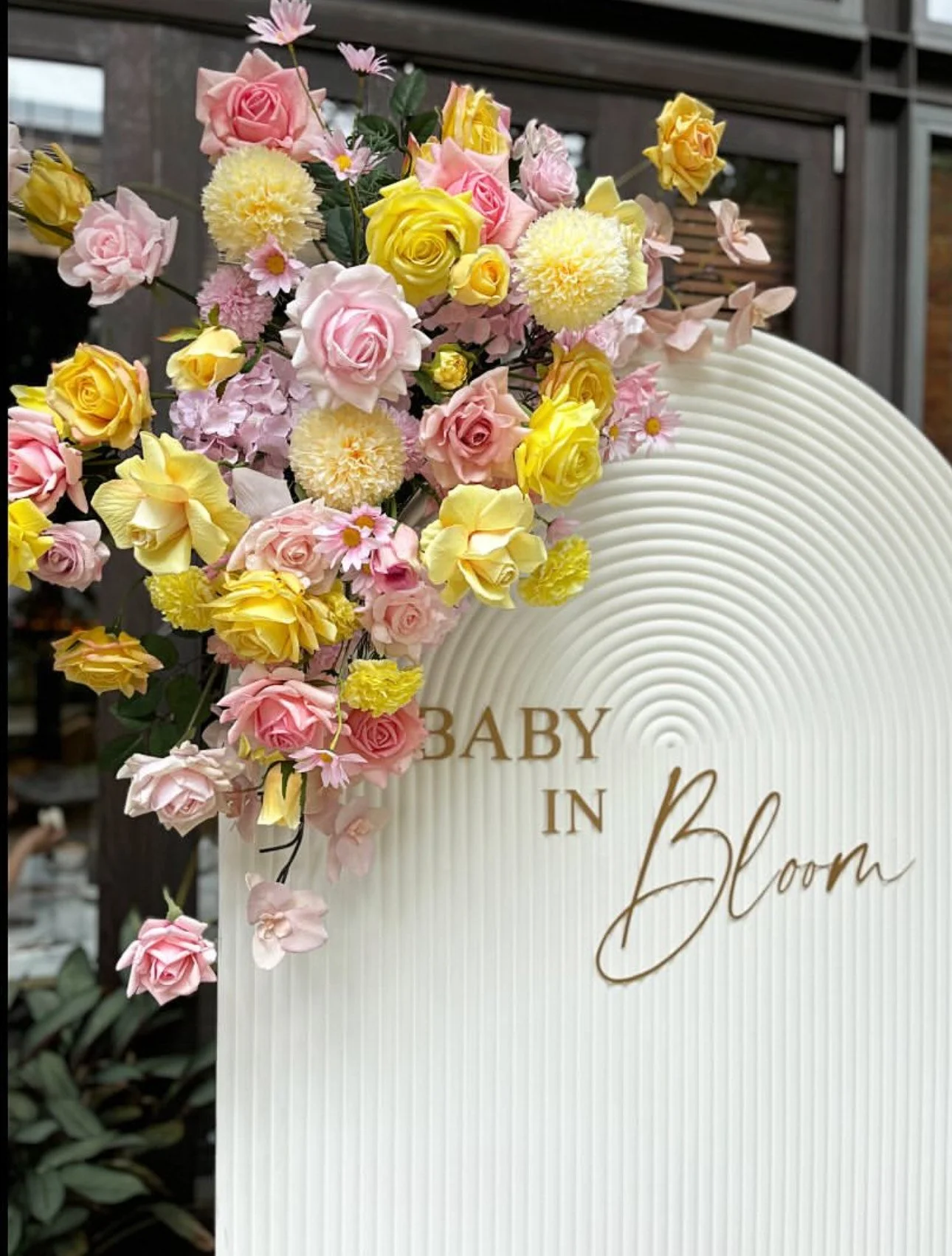 Floral arrangement of pink, yellow, and cream roses, daisies, and other flowers on a white sign that reads "Baby in Bloom"