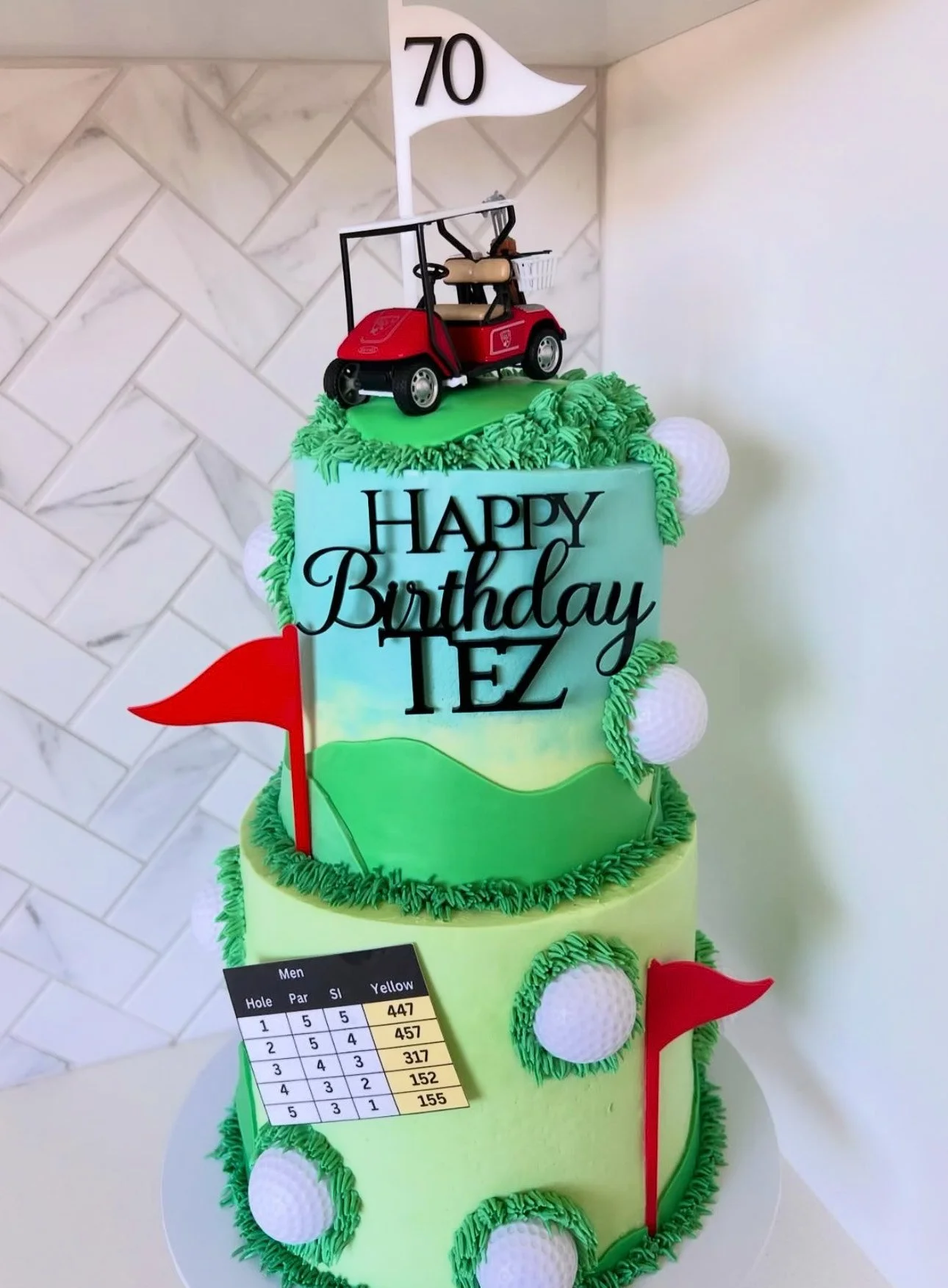 A two-tiered birthday cake decorated with golf theme elements, including golf balls, flags, and a miniature golf cart on top. The top of the cake features a white flag with the number 70, indicating the age celebrated. The cake has personalized writi