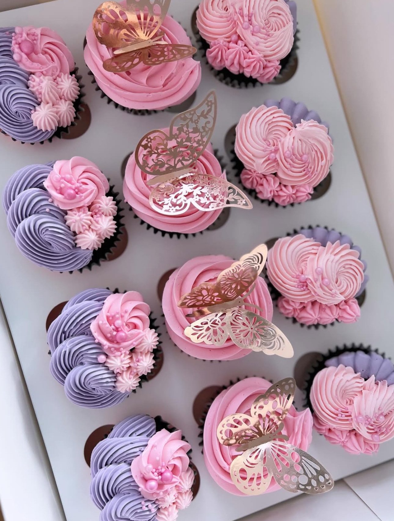 Assorted cupcakes decorated with pink and purple frosting, topped with gold butterfly-shaped edible decorations.