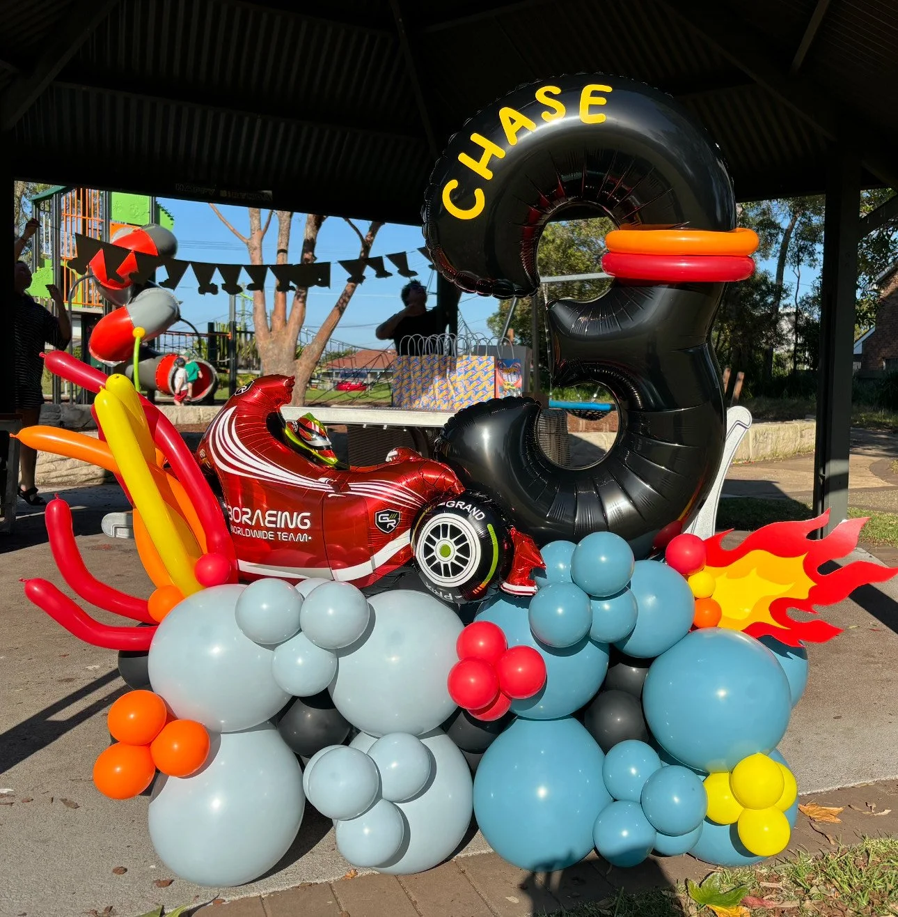 Decorative balloon display featuring a red racing car, a large black tire-shaped balloon with the word 'CHASE' in yellow, flames, clouds of gray and black balloons, and colorful balloon flames. Background shows a park with people, trees, and playground equipment.