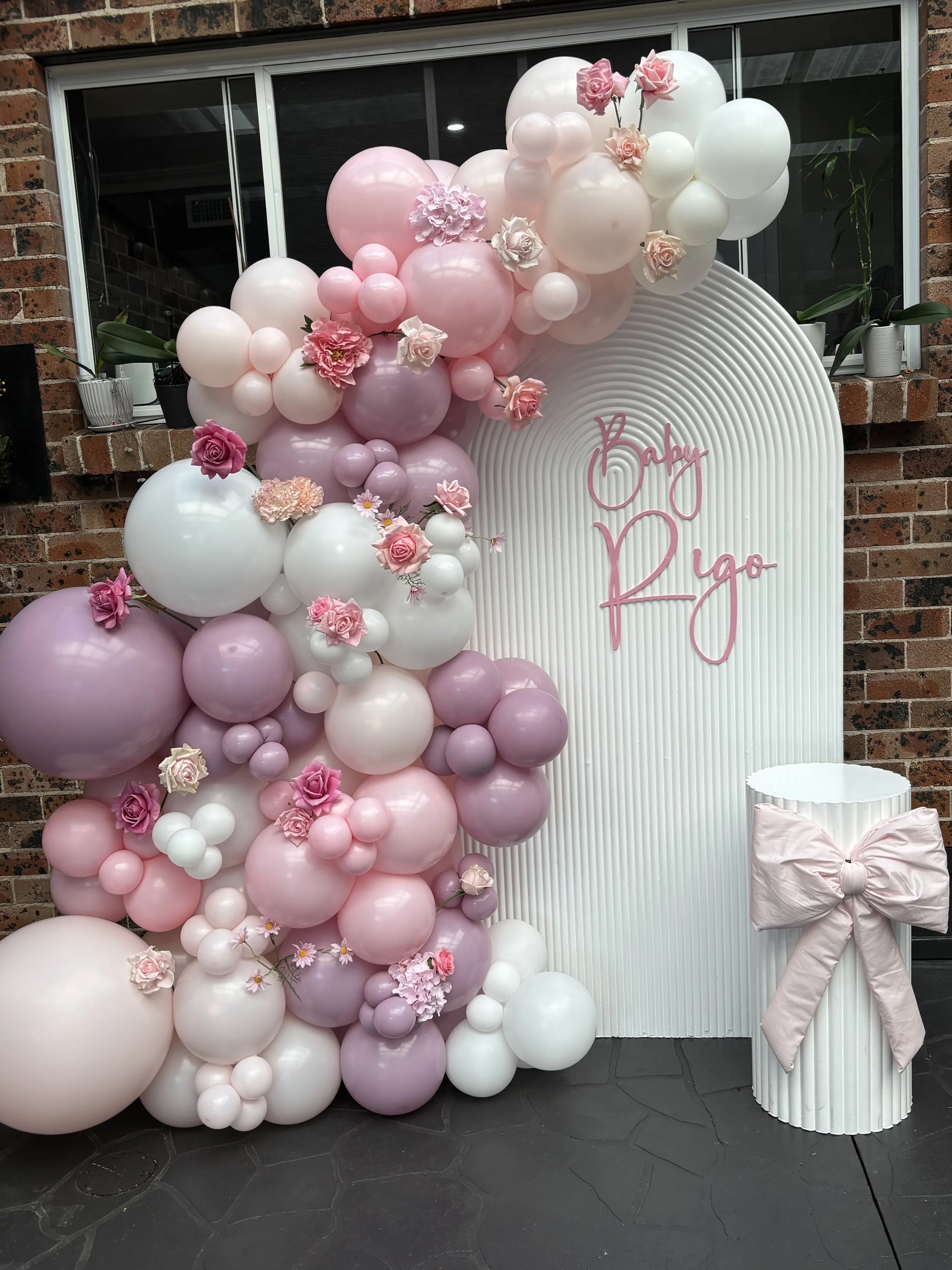 Pink and white balloon display with floral accents in front of a white backdrop with pink lettering that says "Baby Ryo". A cylindrical pedestal with a pink bow is beside the display.