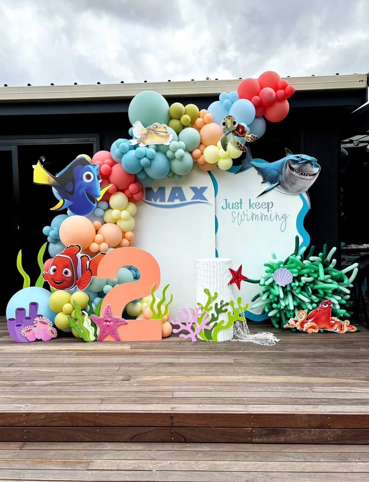 Underwater-themed birthday decoration with balloons, toy characters from Finding Nemo and Finding Dory, a large pink number two, coral, starfish, and seaweed, on a wooden deck.